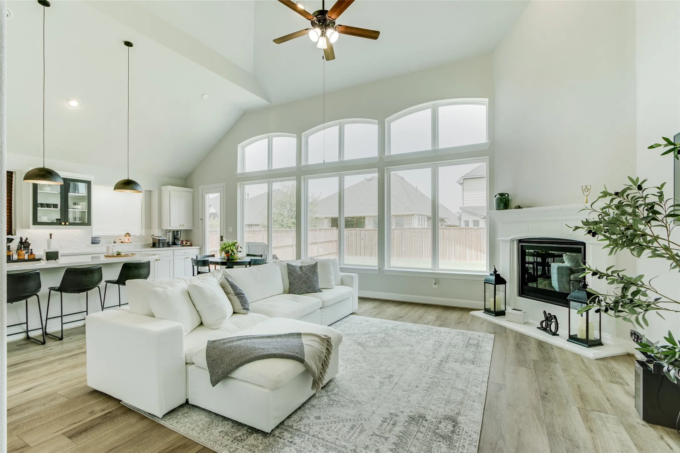 Vaulted ceiling with a ceiling fan, expansive wall of windows, integrated fireplace with a mantel, wood-finish flooring, and a kitchen island with pendant lighting