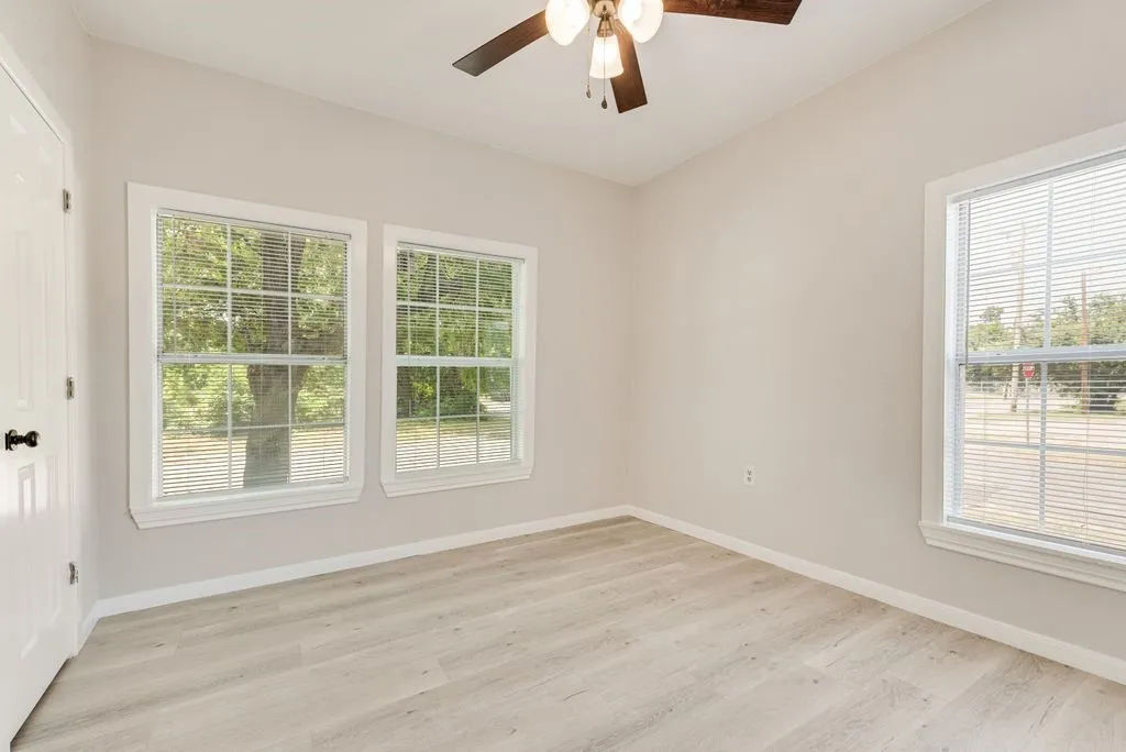 Light wood-finish flooring, white trim, multiple windows with blinds, a white paneled door, and a ceiling fan with integrated lighting