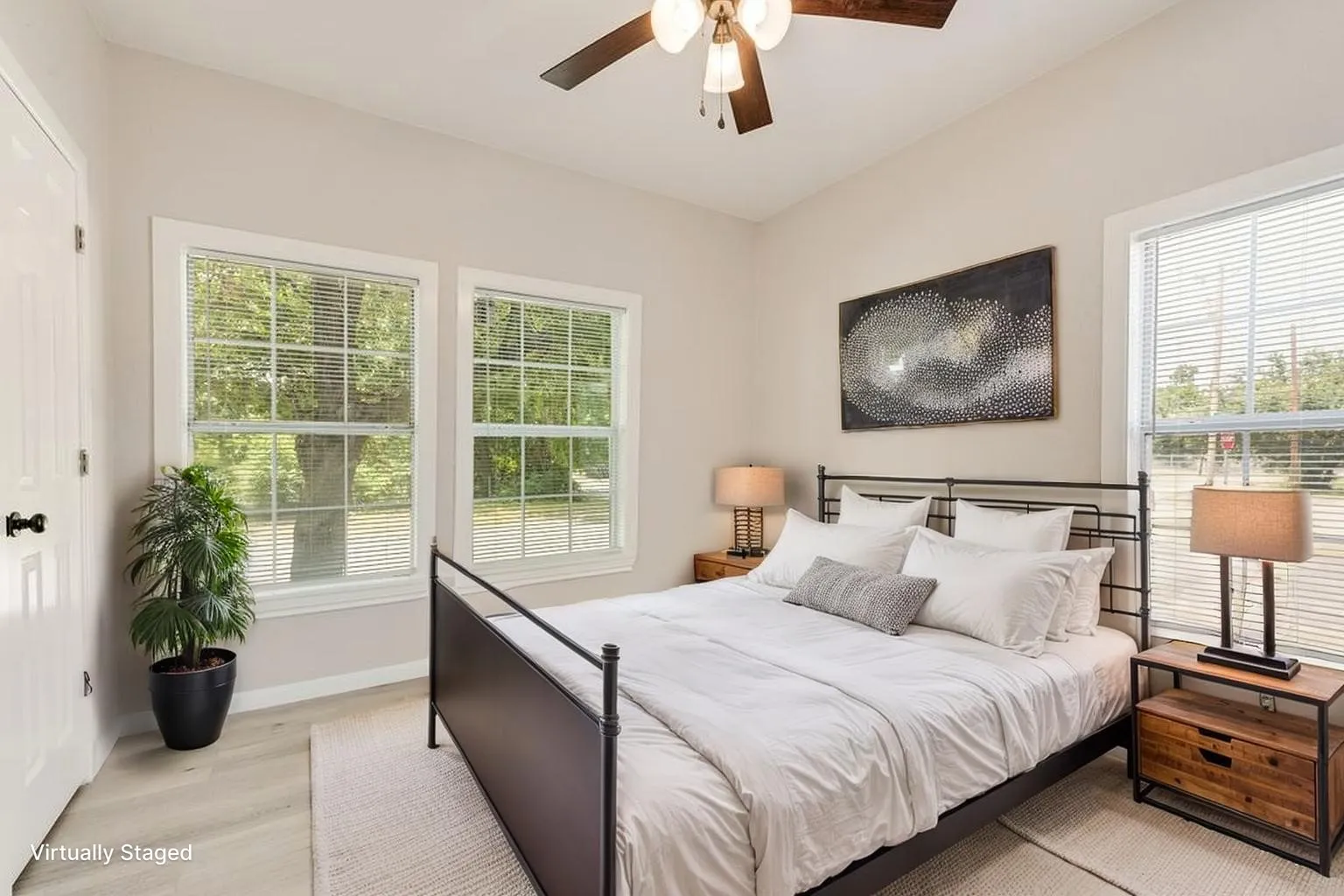 Virtually staged Bright, light-filled bedroom featuring multiple large windows with plantation shutters and transom panes, a ceiling fan with integrated lighting, wood-finish flooring, and a metal bed frame with horizontal slat detailing