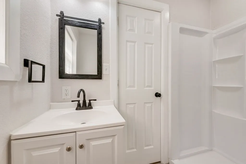 Clean, bright bathroom featuring a white vanity with dual cabinet storage, white subway tile backsplash, brushed bronze faucet and hardware, framed black-trimmed mirror, and white six-panel door entry