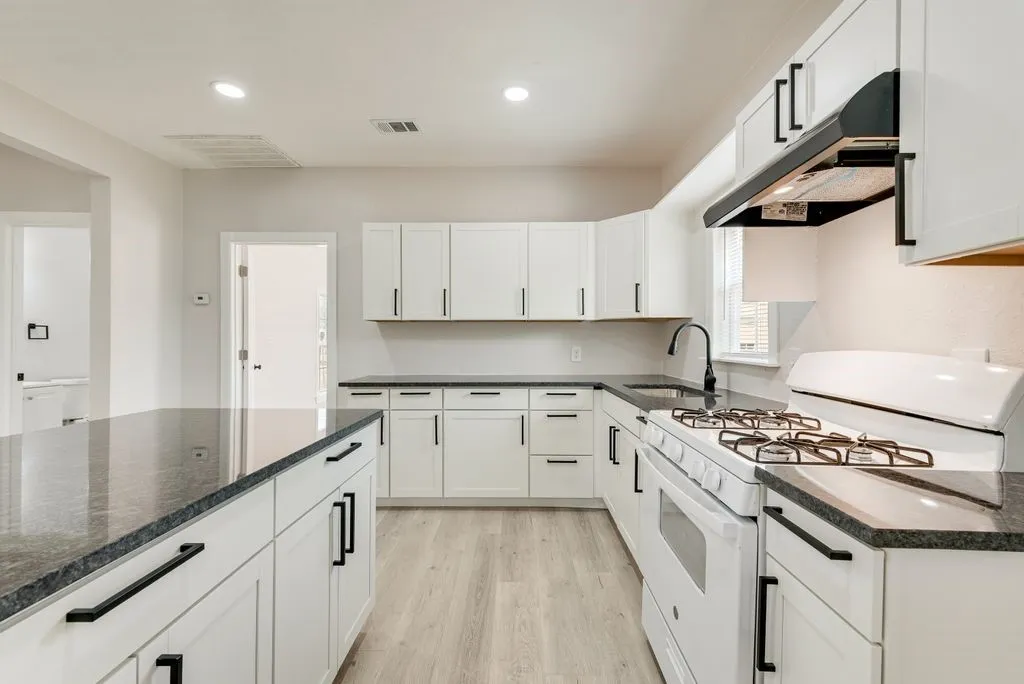 Modern kitchen featuring white cabinetry with matte black hardware, gas cooktop with stainless steel hood, stainless steel appliances, dark wood-finish flooring, and dark stone countertops throughout the galley-style layout with recessed lighting and pendant fixtures