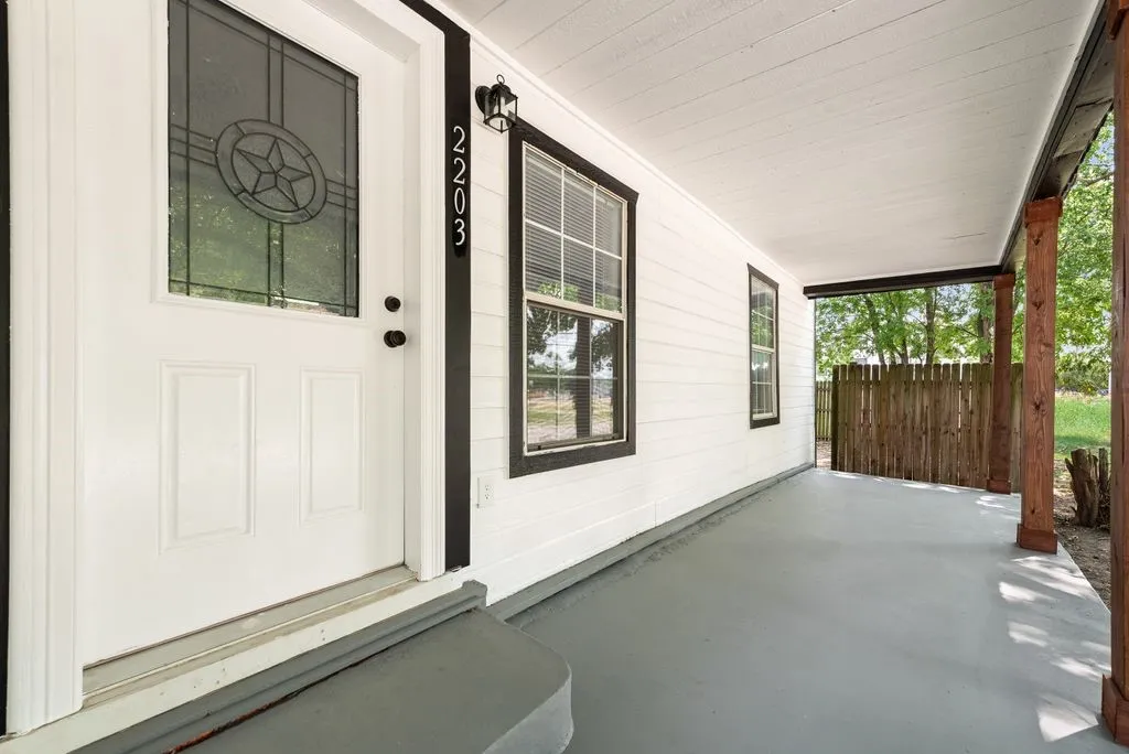Covered front porch featuring a concrete floor, white-painted exterior walls with black-trimmed windows and entry door, decorative transom window detail, and mature tree backdrop with natural lighting throughout