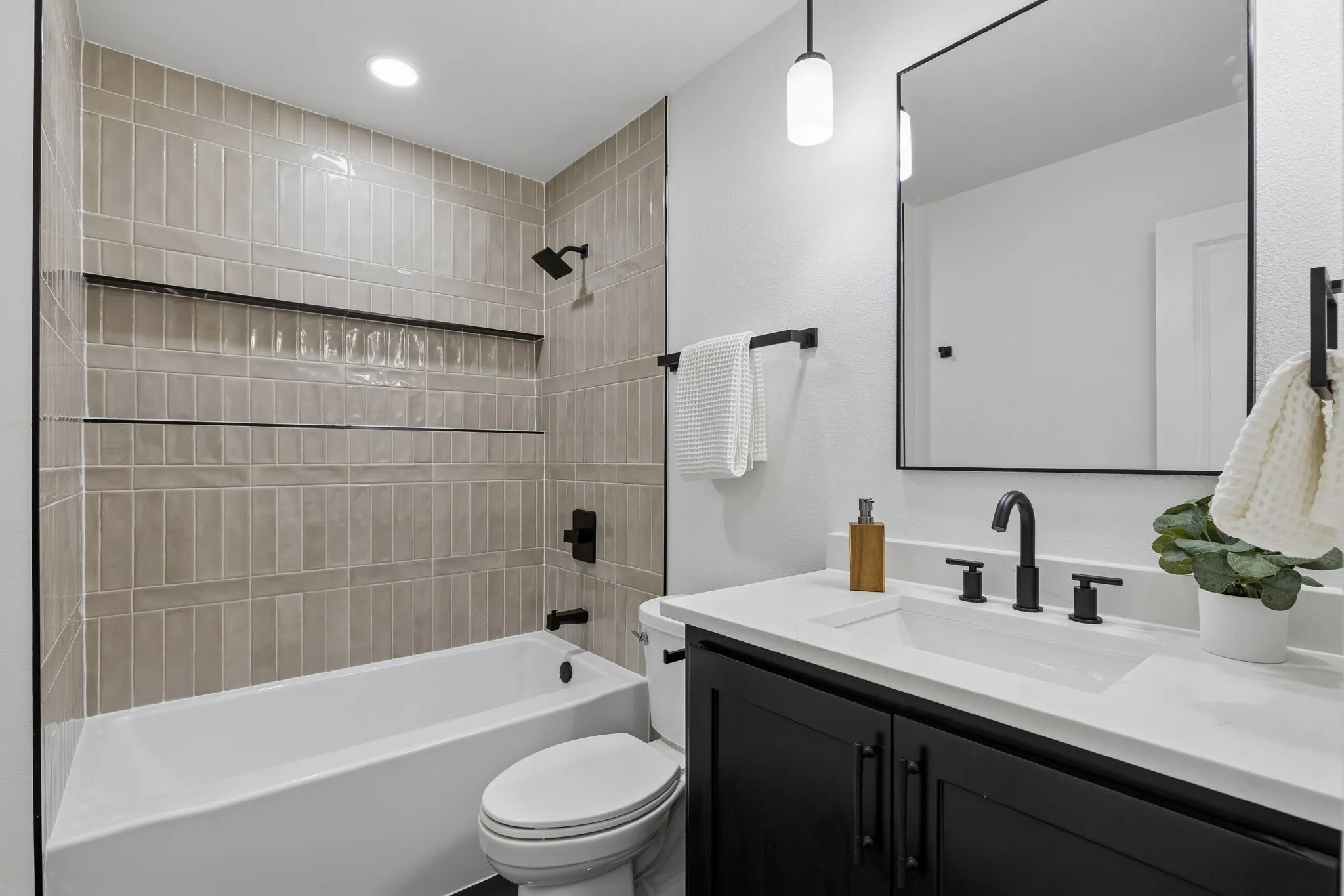 Bathtub and shower combination with vertical tile surround and built-in shelving, black vanity with an integrated sink and white countertop, black framed mirror, and matte black fixtures