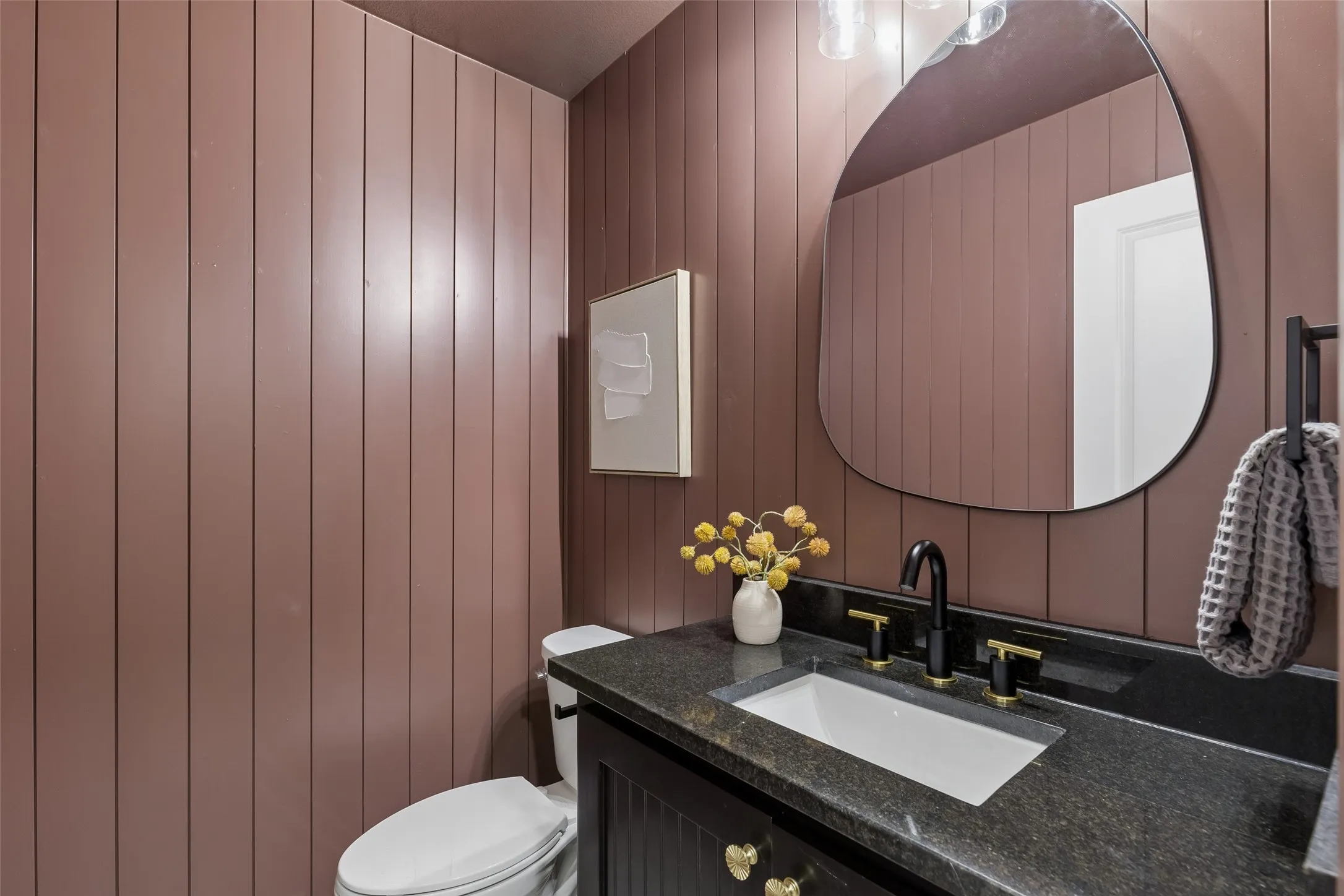 Powder room featuring vertical shiplap paneling in warm mauve tones, a curved organic mirror with integrated lighting, dark stone countertop with undermount sink basin, matte black faucet hardware, white porcelain toilet, and recessed wall-mounted medicine cabinet