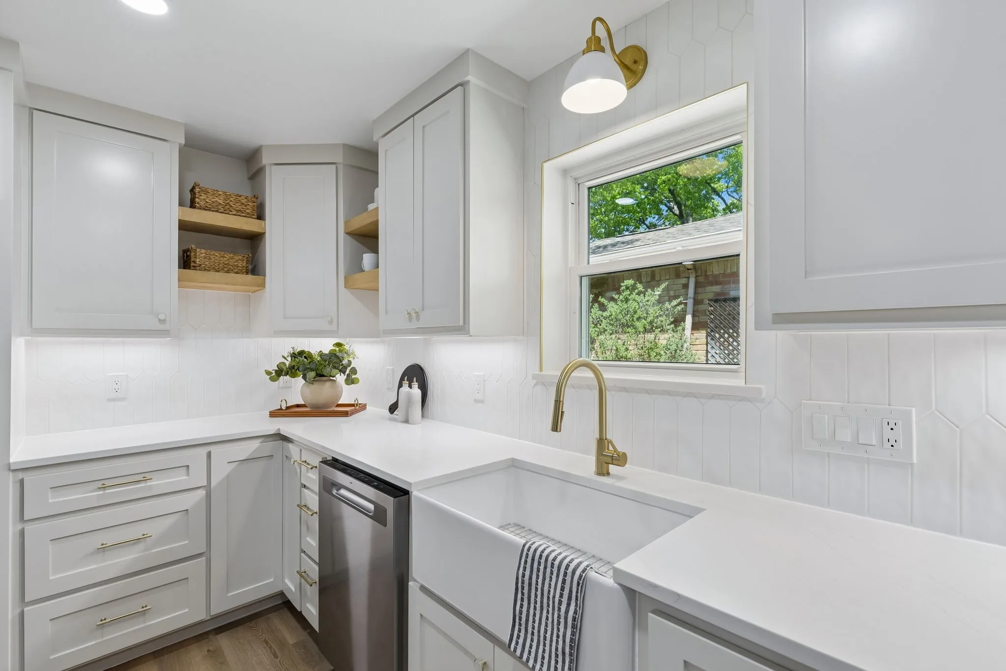 White cabinetry with gold-tone hardware, white countertops, built-in stainless steel dishwasher, and a white farmhouse sink with a gold-tone faucet