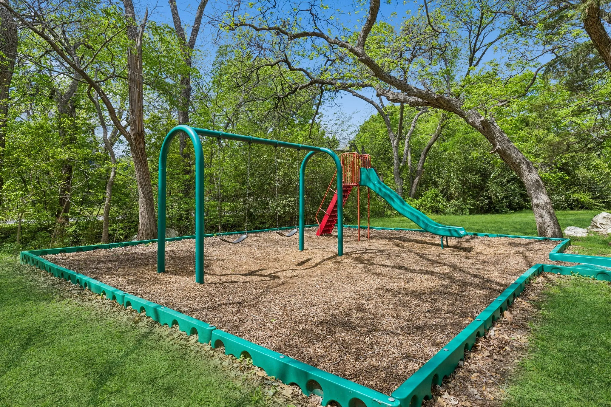 Outdoor play area featuring a swing set, slide, and wood chip ground cover, bordered by green edging
