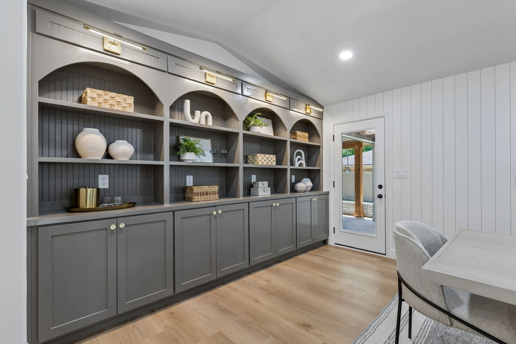 Vaulted ceiling room featuring extensive built-in shelving with arched details, lower cabinetry with brass hardware, wood-finish flooring, and a glass-paneled door