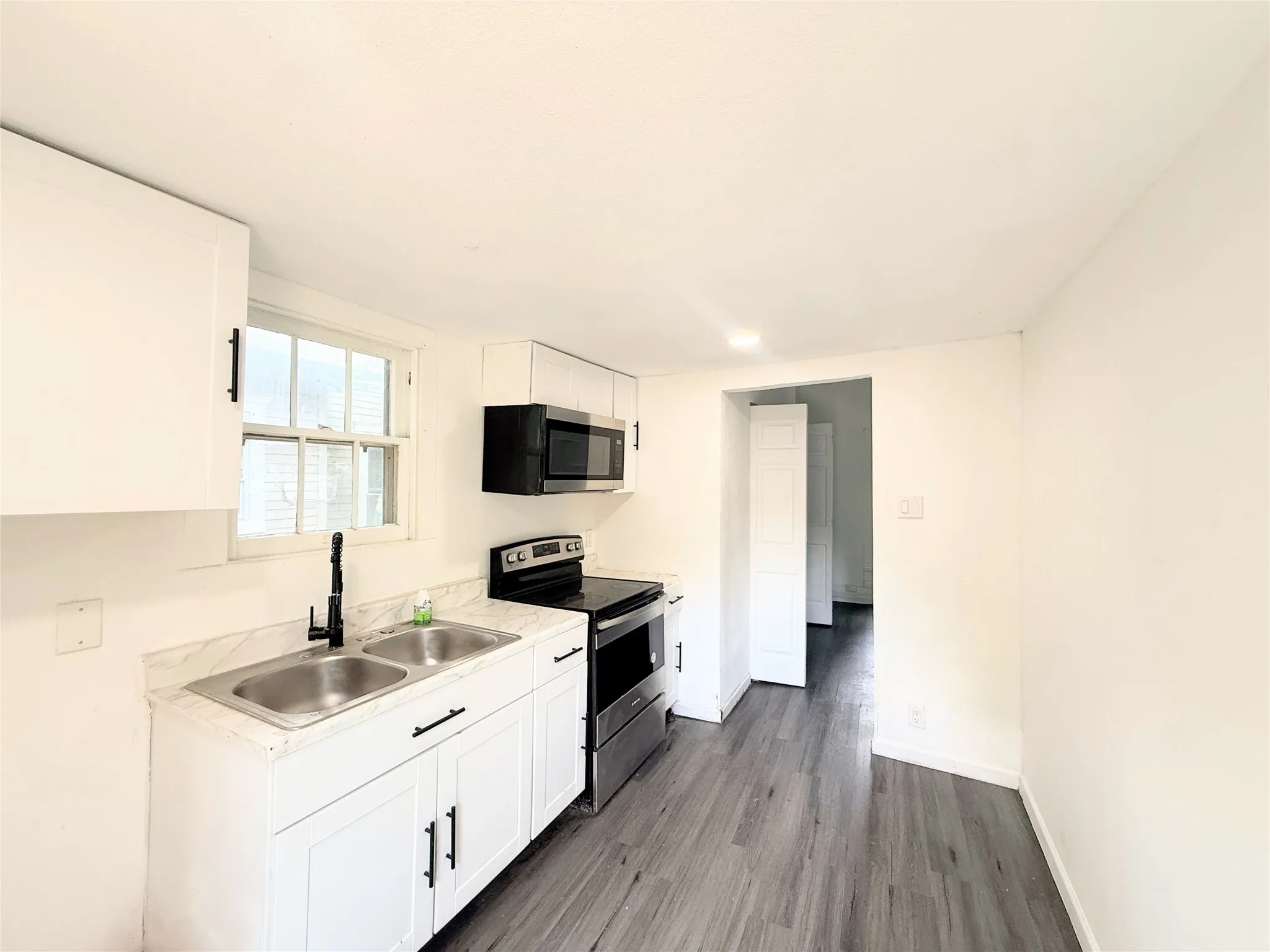 Modern galley kitchen featuring white cabinetry with black hardware, stainless steel appliances including a black electric range and microwave, a stainless steel sink with single-handle faucet, wood-finish flooring throughout, and abundant natural light from a large window