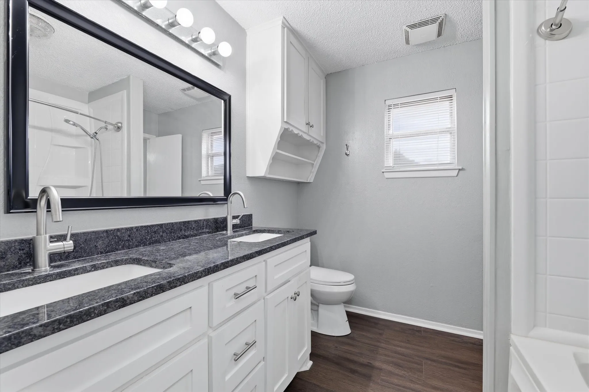 Dual vanity with undermount sinks, stone-finish countertop, white cabinetry with silver-tone hardware, wood-finish flooring, and a shower-tub combination