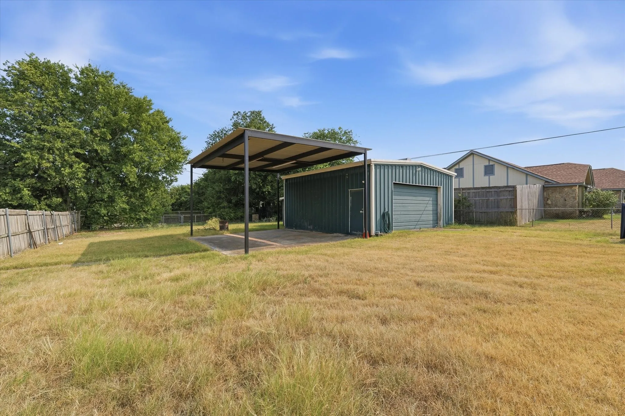 Expansive grounds featuring mature shade trees and open pasture, complemented by a detached metal storage structure with covered carport, additional outbuildings, and perimeter fencing