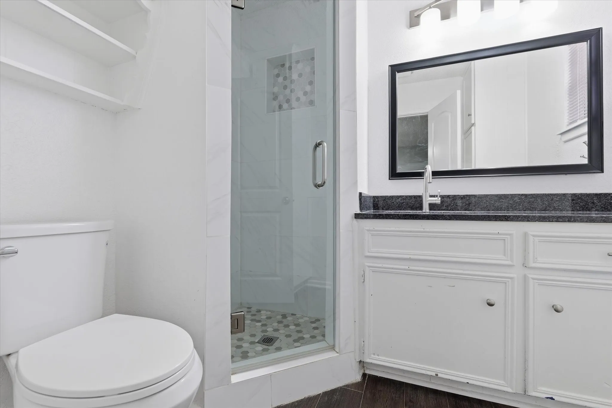 Spa-inspired bathroom featuring a frameless glass shower enclosure with mosaic tile flooring, dual vanity with white cabinetry and dark countertop, large framed mirror, and contemporary hardware throughout