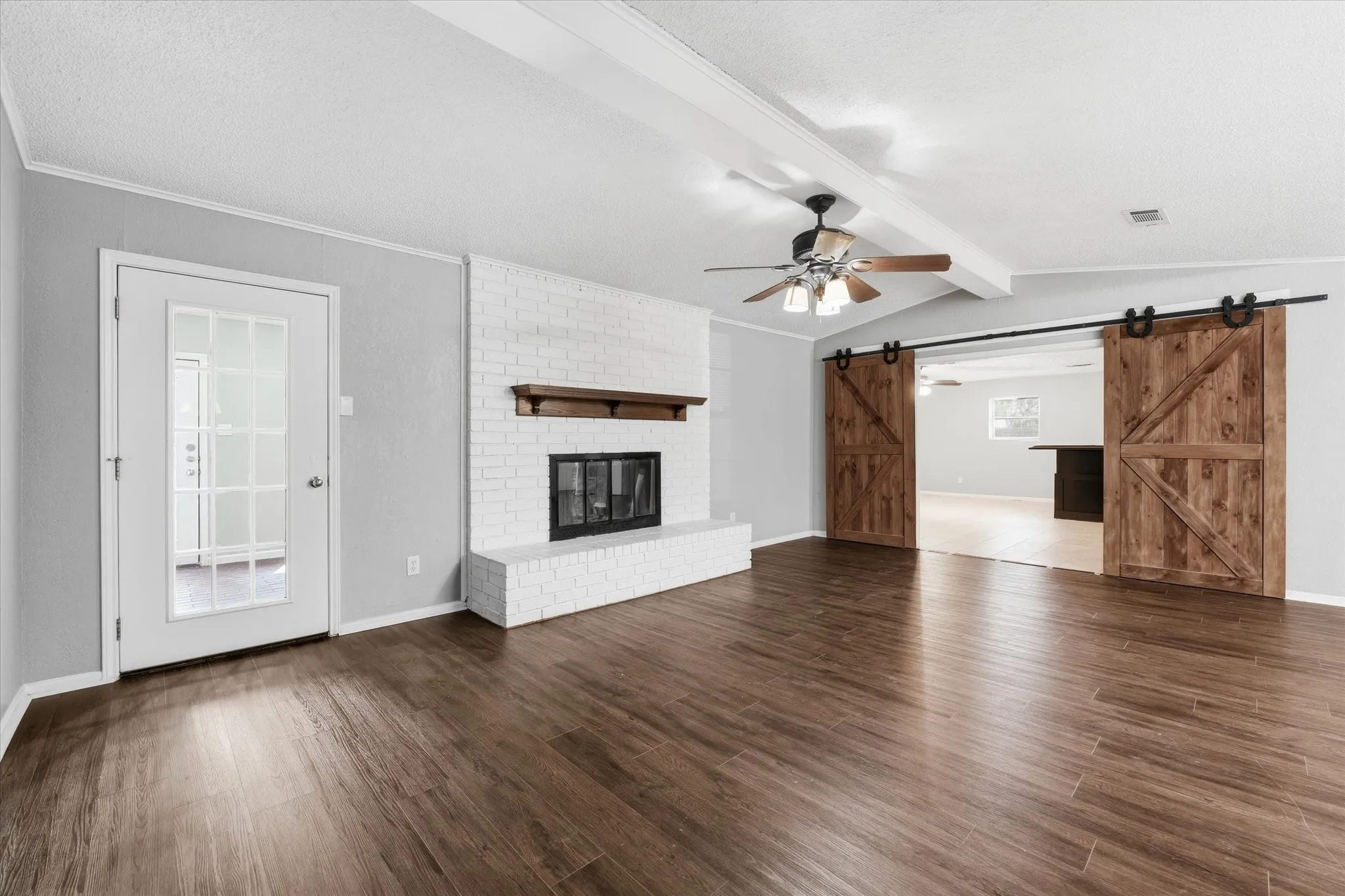 Spacious living area featuring wood-finish flooring, a white brick fireplace with a wood mantle, and exposed ceiling beams