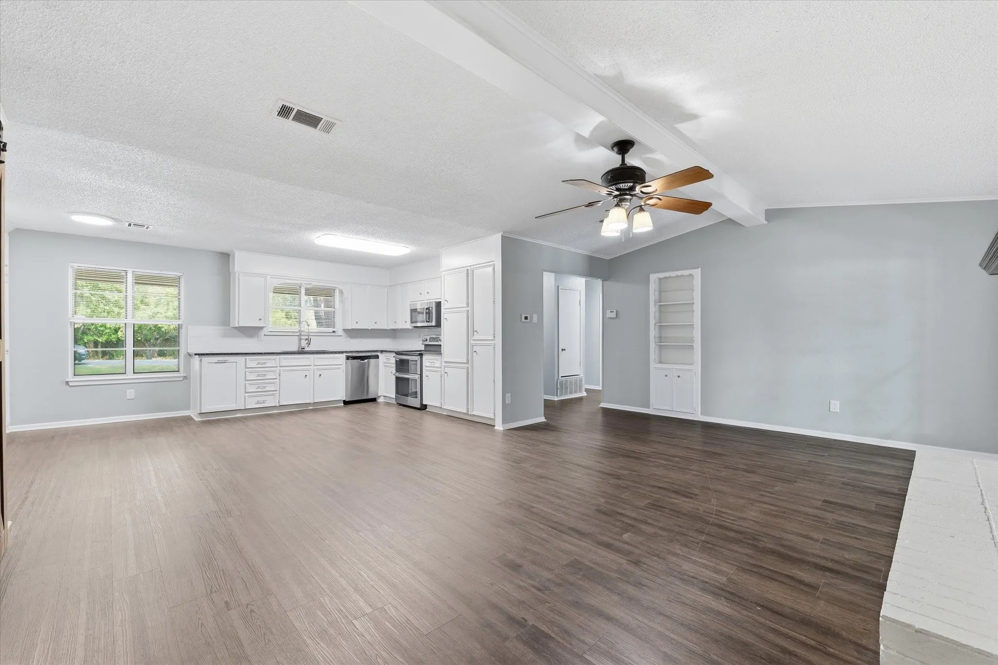 Open-concept living space featuring warm wood-finish flooring throughout, white cabinetry with stainless steel appliances, recessed ceiling lighting, and a ceiling fan with integrated light fixture