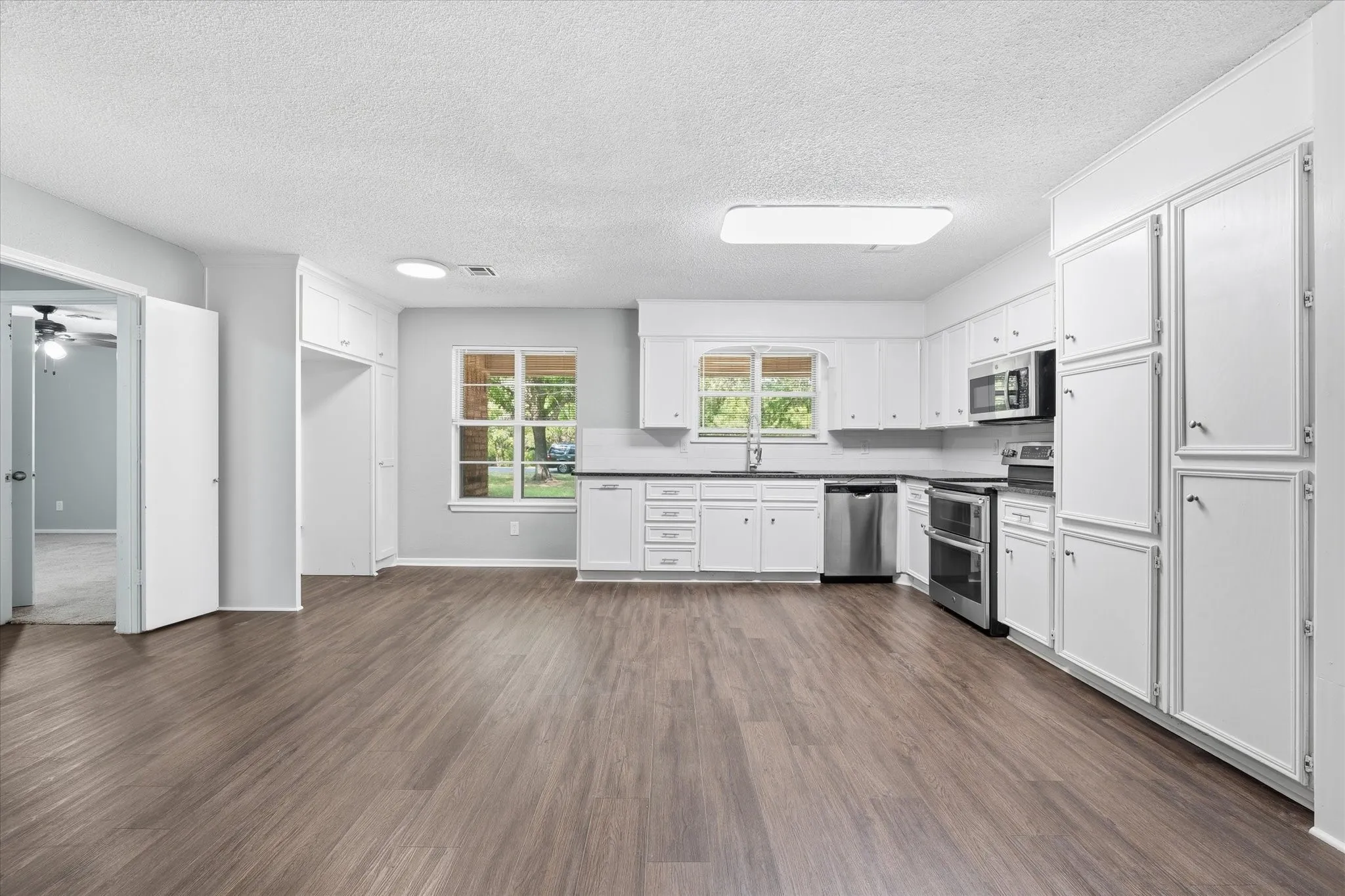 Spacious kitchen featuring white cabinetry, stainless steel appliances, wood-finish flooring throughout, abundant natural light from multiple windows, and a generous open floor plan ideal for entertaining