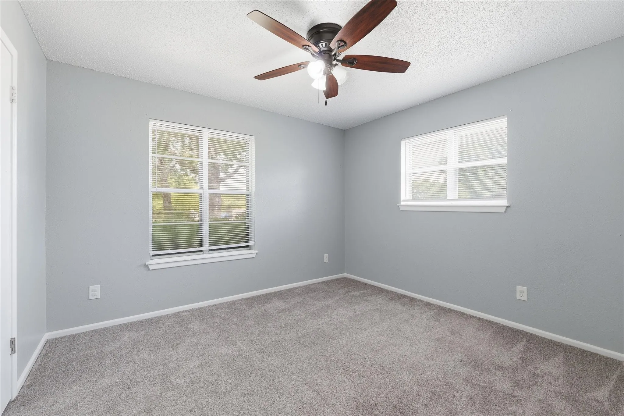 Neutral gray walls, two windows with blinds, gray carpet, and a ceiling fan with light fixture