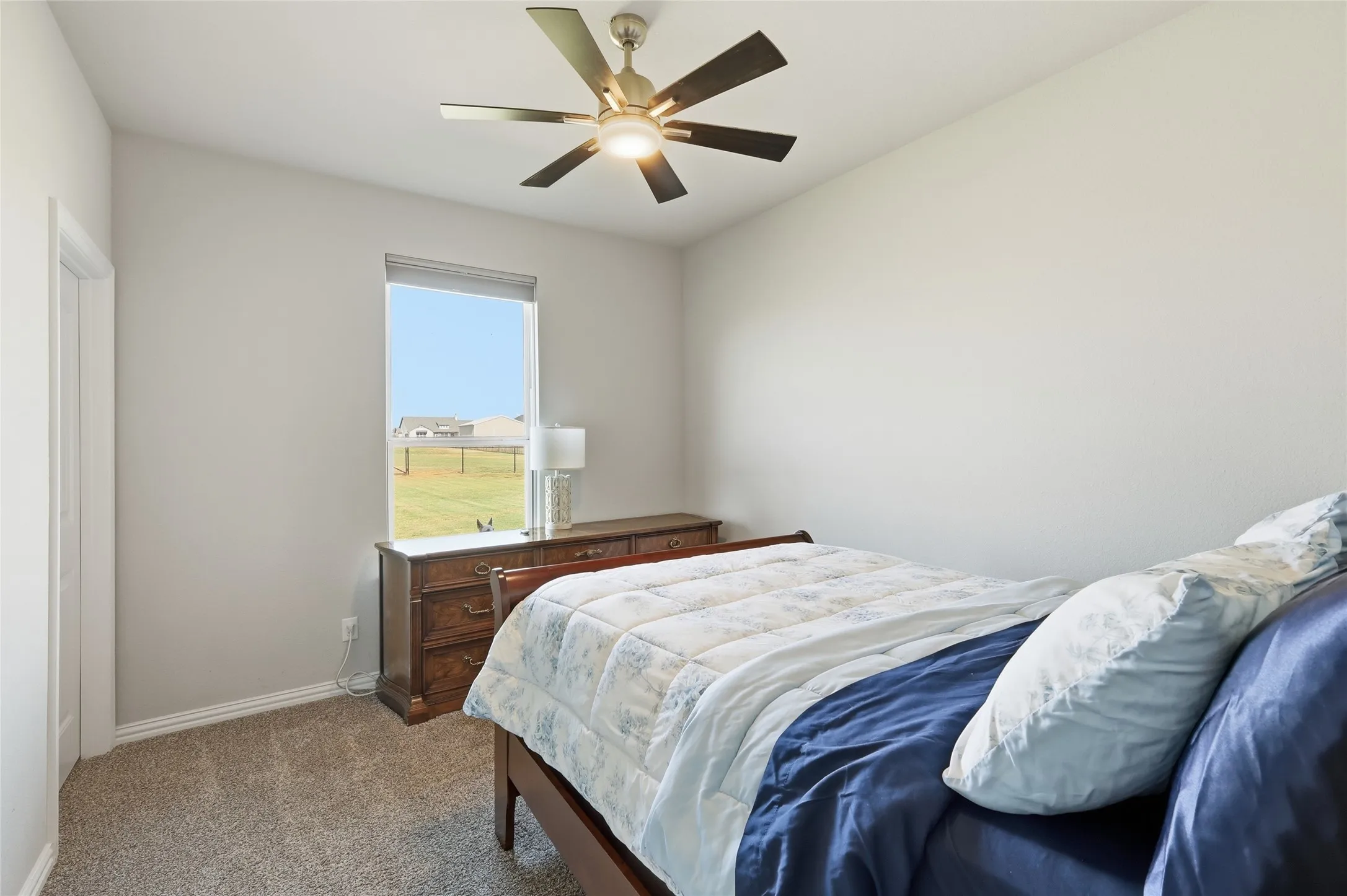 Bright secondary bedroom featuring carpeted flooring, wood-finish dresser, ceiling fan with integrated lighting, and large window with natural light and pastoral scenery