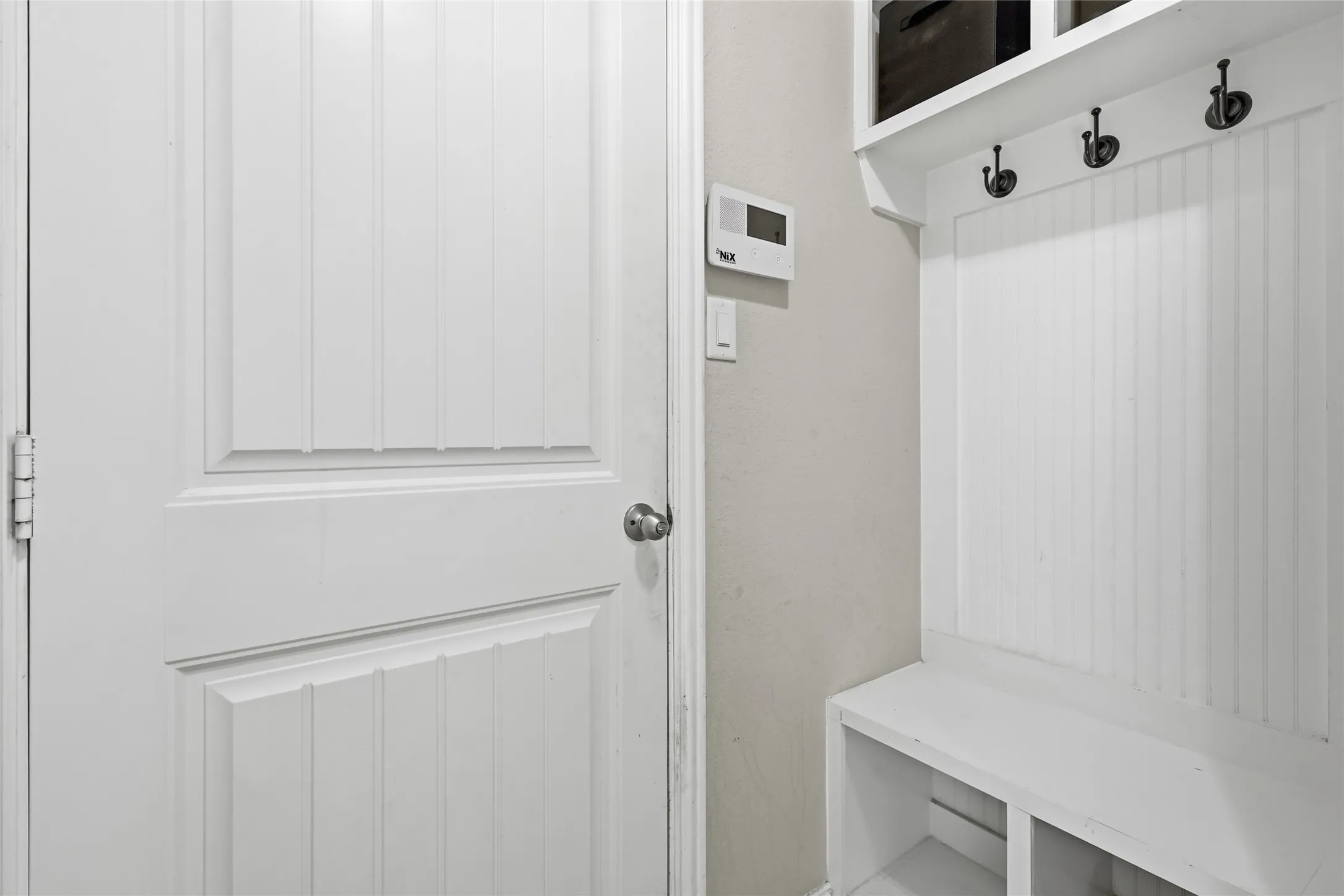 Bright mudroom entry featuring white shaker-style cabinetry with built-in bench seating, overhead hooks for coat storage, and white vertical paneling throughout
