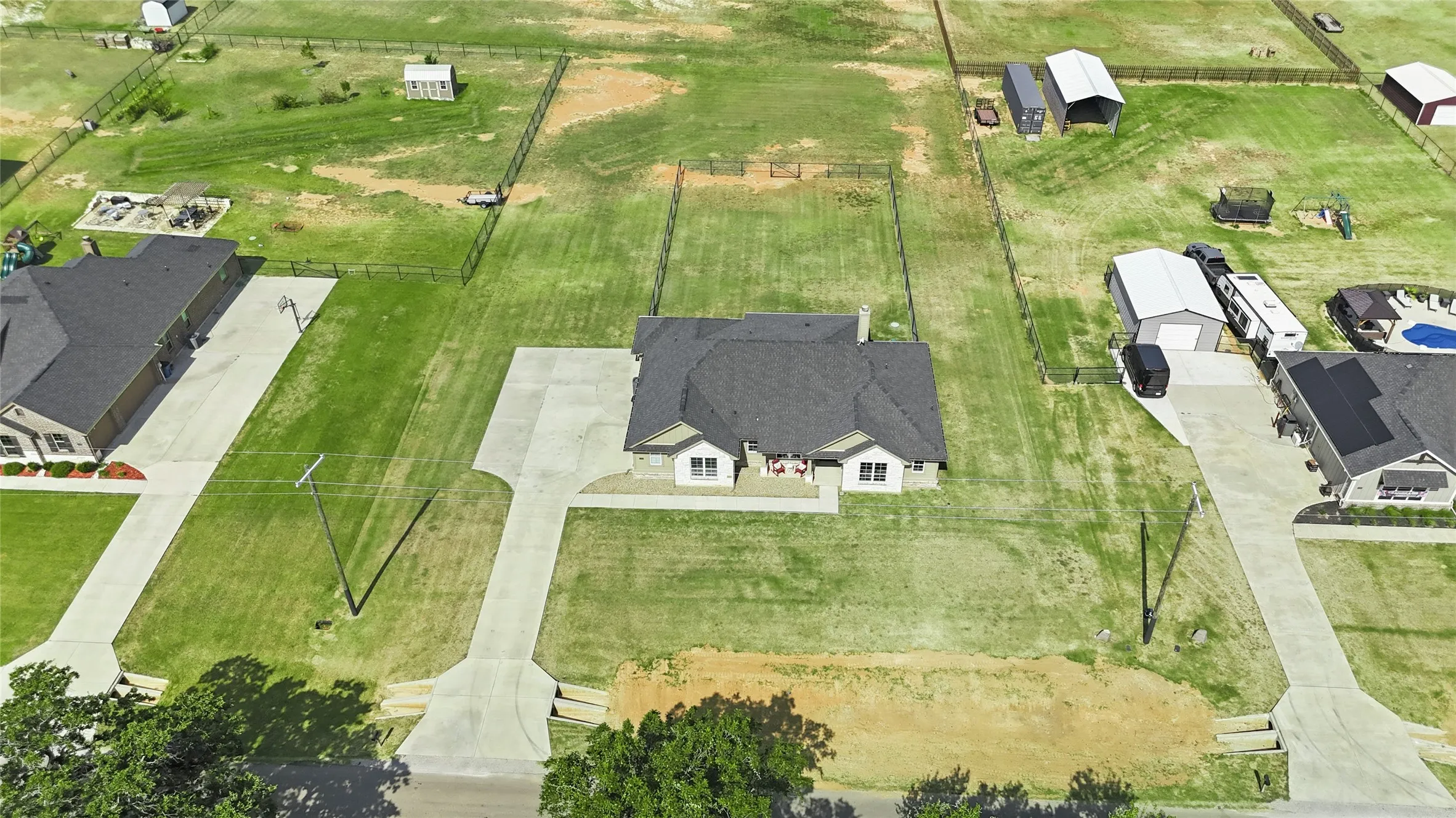 Aerial view showcasing this residential property situated on a spacious lot with manicured lawn, concrete driveway, and a single-story home featuring a dark-shingled roof