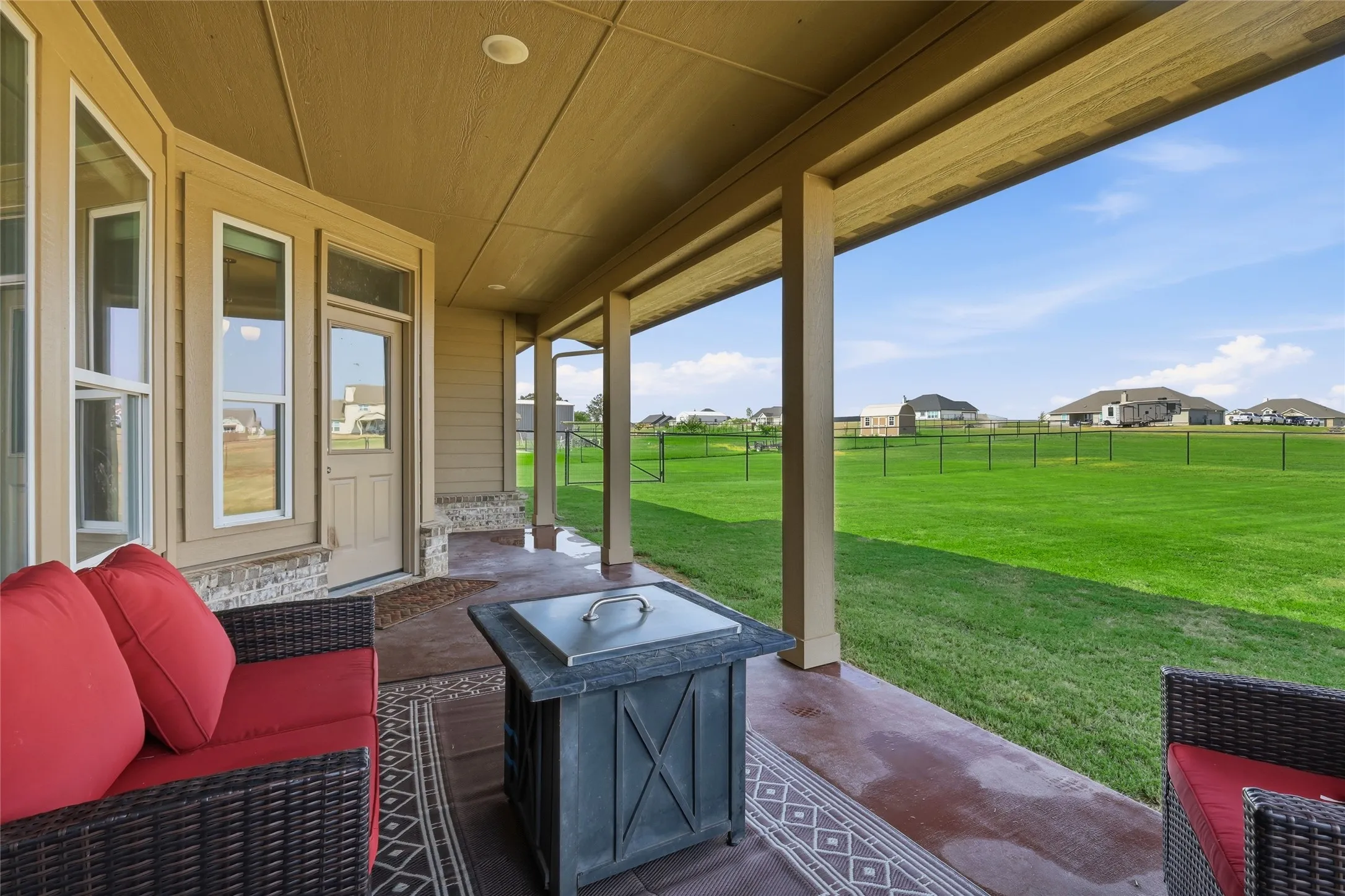 Expansive covered porch with wood-finish ceiling and beam details, featuring a fire pit with stone surround, wicker seating with red cushions, and direct access to manicured grounds