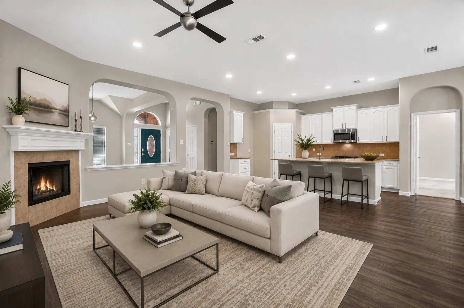 Virtually staged Integrated kitchen and living area, featuring a decorative fireplace with a mantel, dark wood-finish flooring, white cabinetry, and a ceiling fan
