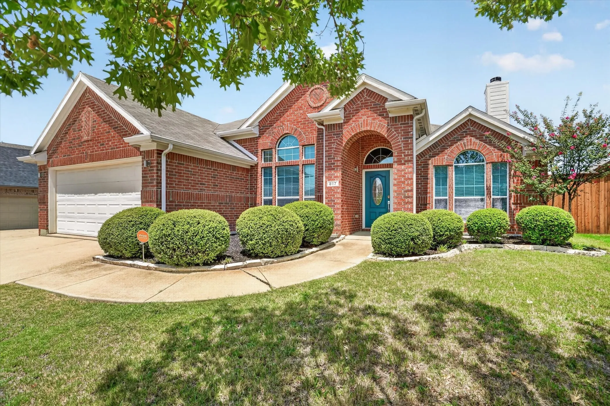 Charming brick home featuring an arched entryway with transom window, dual curved shrub landscaping, manicured lawn, two-car garage with white panel door, and professional concrete driveway and walkway