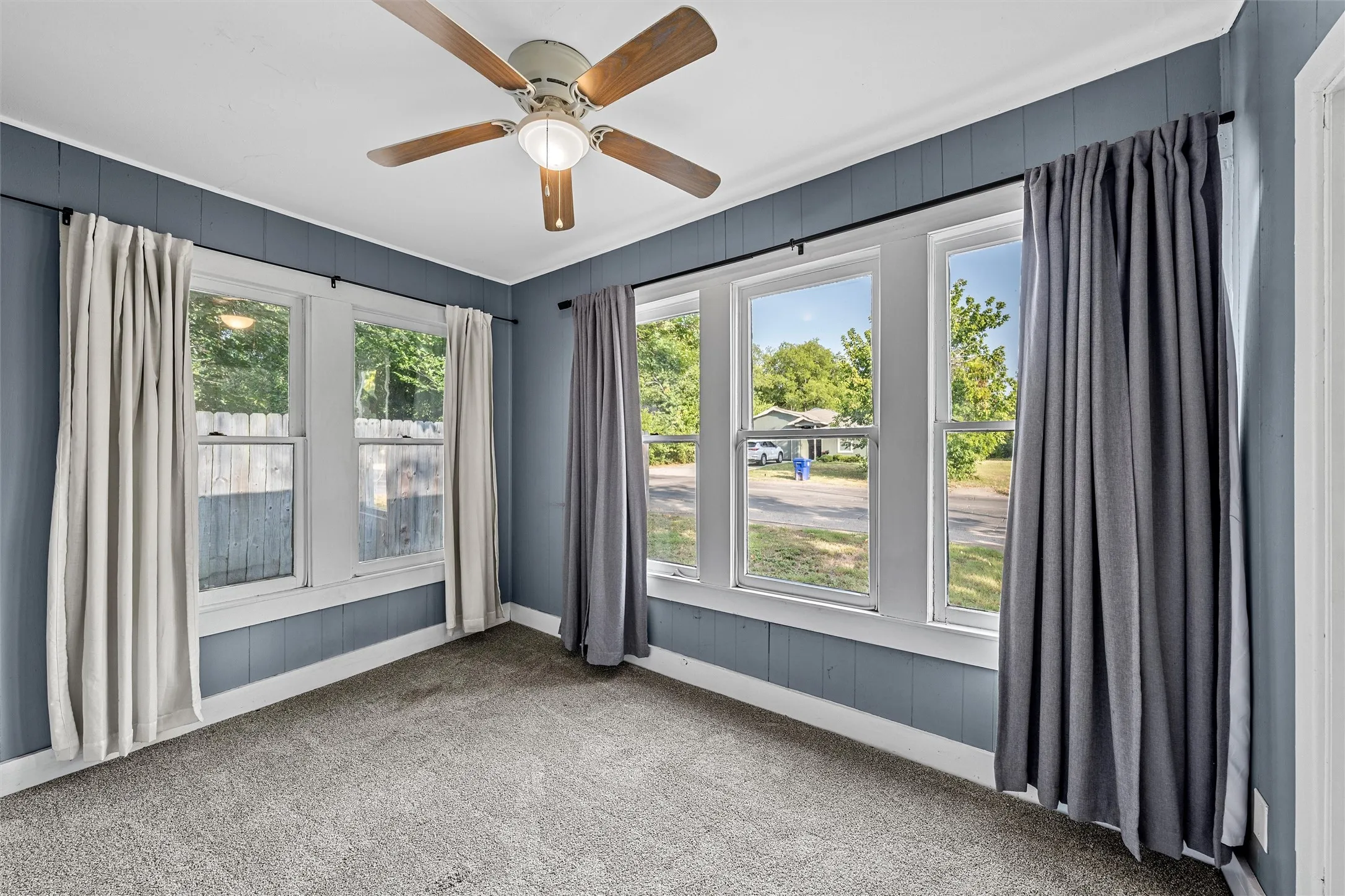 Bright corner sunroom showcasing multiple large windows with natural light, neutral carpet flooring, ceiling fan with integrated lighting, and gray shiplap accent walls framing expansive outdoor sightlines