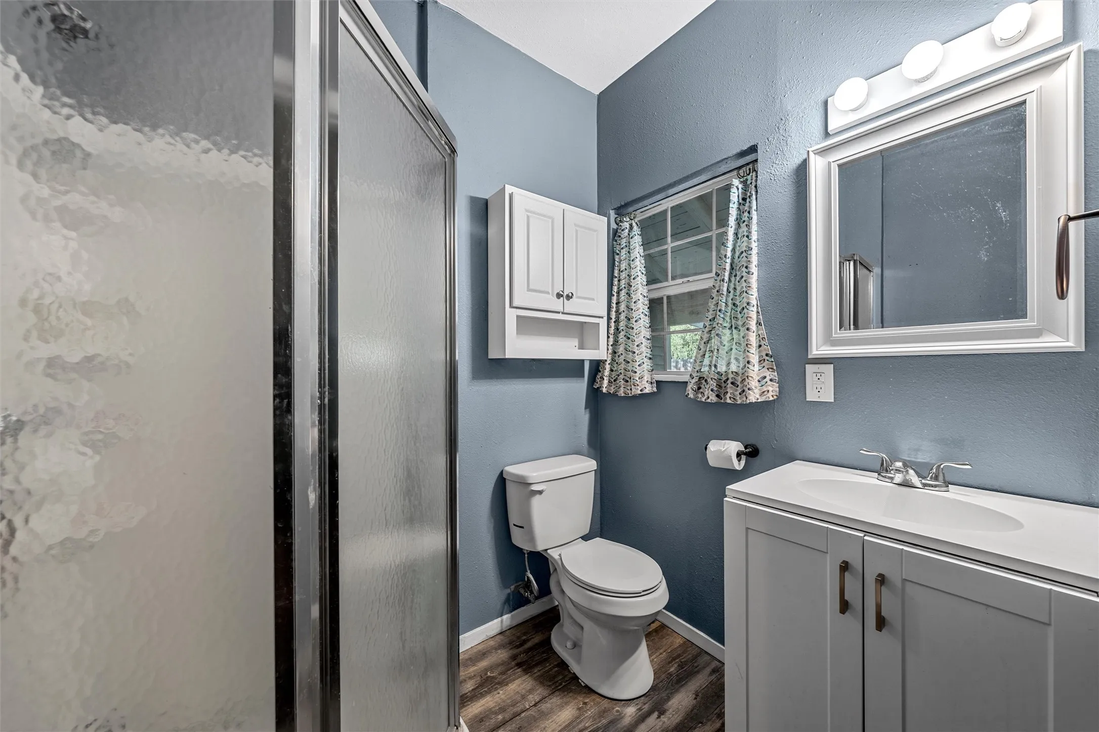 Shower enclosure with textured glass, vanity with integrated sink, wood-finish flooring, wall-mounted medicine cabinet, and framed mirror