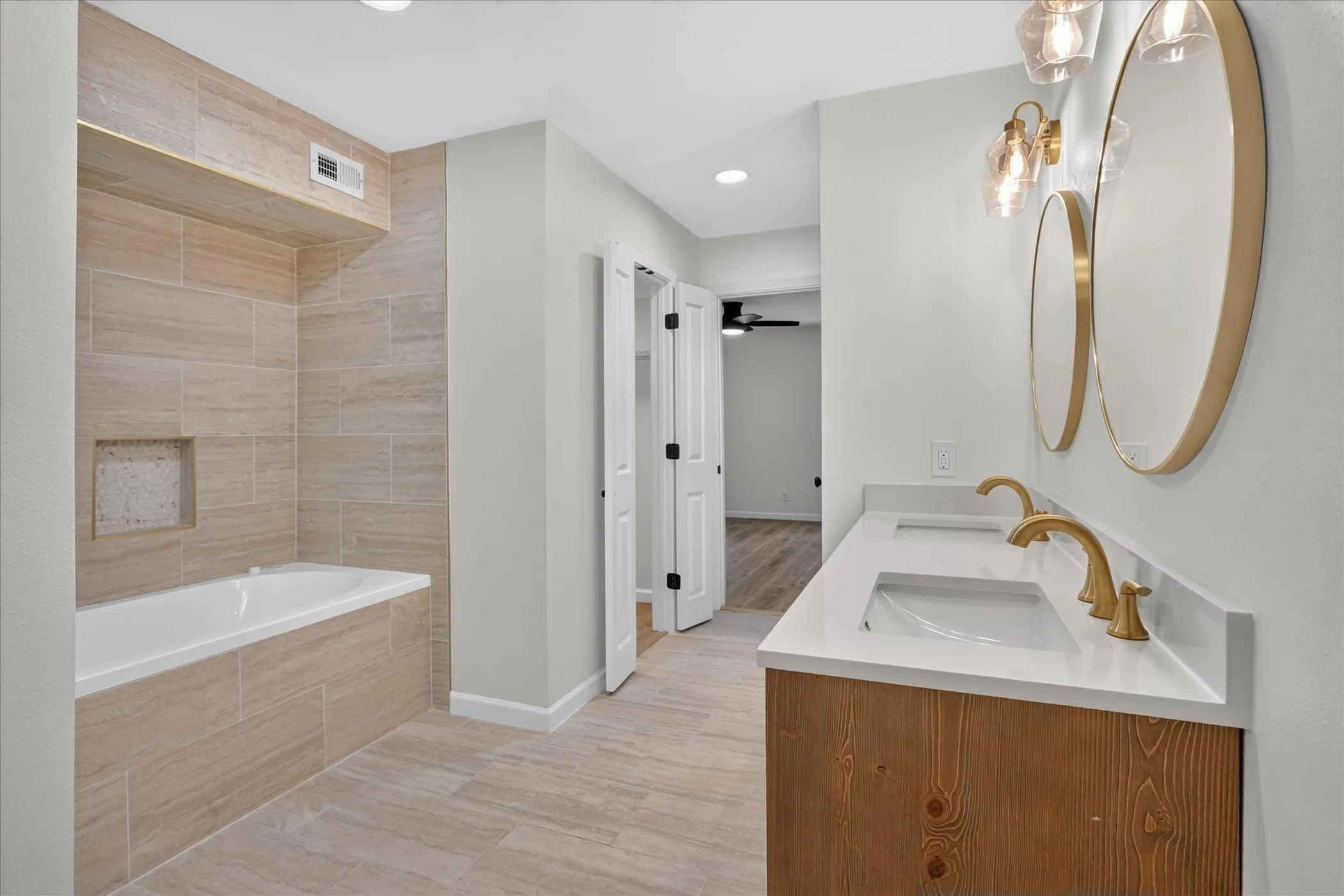 Primary bathroom at 6020 E University Blvd Unit 117 Dallas Soaking tub with full large-format tile surround, brushed gold faucet, fluted wood vanity, and walk-in closet access.