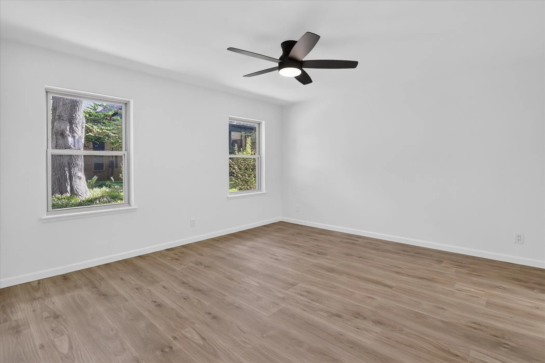 Light-filled bedroom at 6020 E University Blvd Unit 117 Dallas. New LVP flooring, ceiling fan, and dual windows with mature tree views in gated East Dallas community.