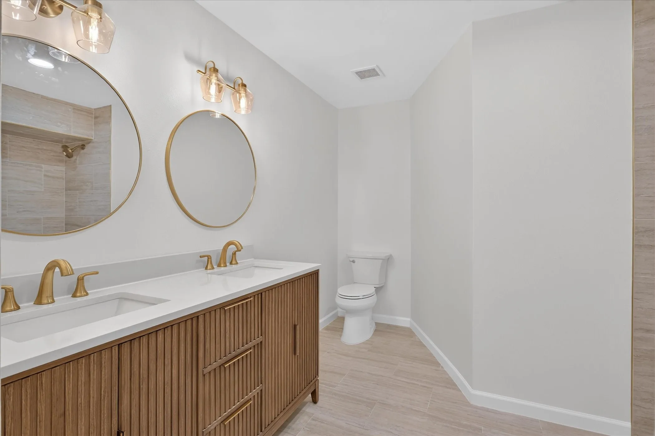 Renovated bathroom at 6020 E University Blvd Unit 117. Dual undermount sinks, fluted wood vanity cabinet, brushed gold fixtures, round gold-framed mirrors, and walk-in shower.