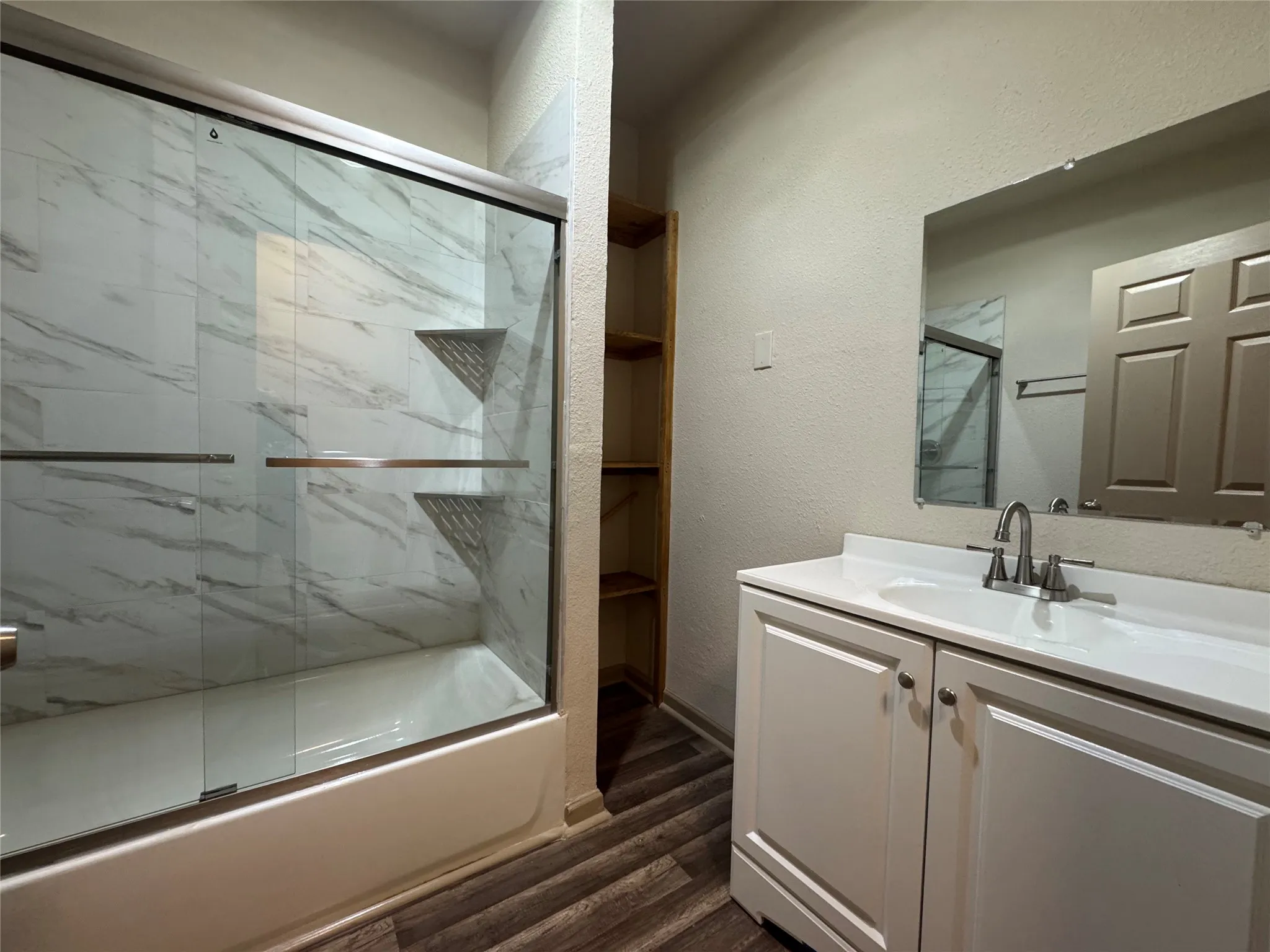 Bathroom featuring a tub/shower combination with sliding glass doors, a white vanity with an integrated sink, built-in shelving, and wood-finish flooring