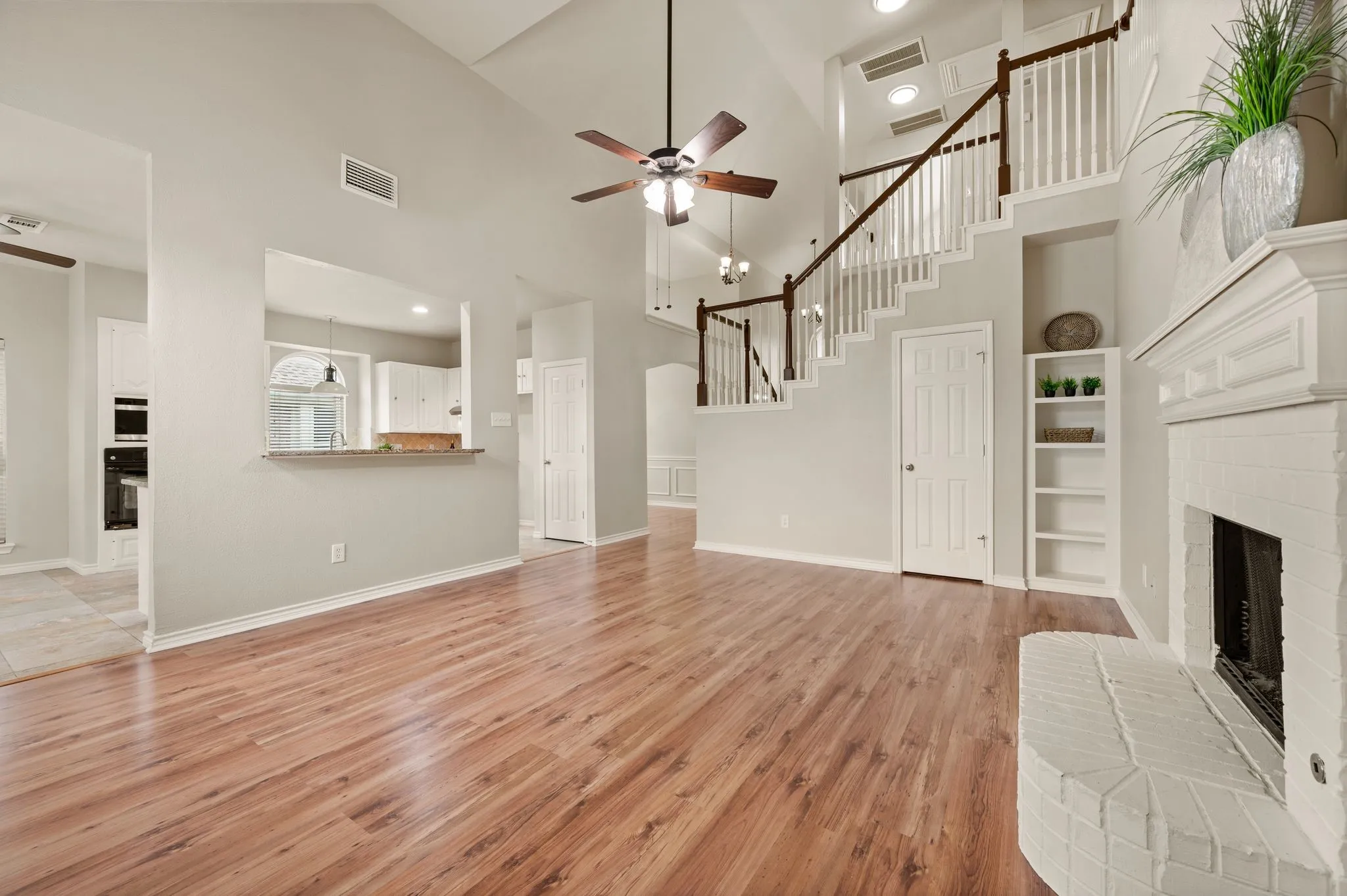 Spacious living area featuring wood-finish flooring, a white brick fireplace with mantel, and a multi-level staircase with white balusters and dark wood handrails