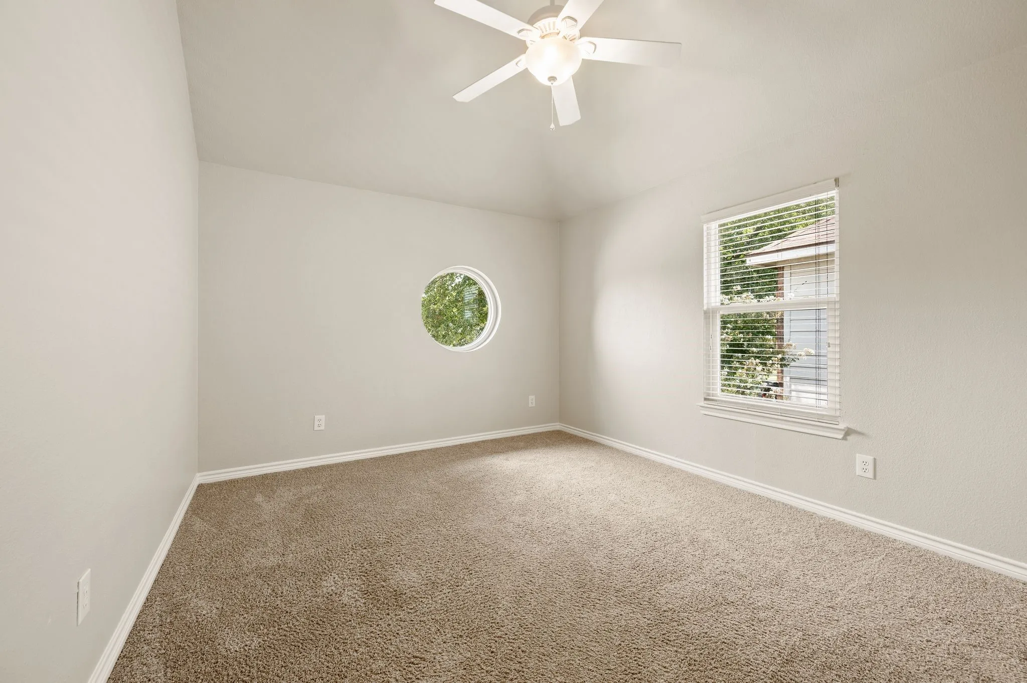 Carpeted bedroom featuring a distinctive round window, an additional rectangular window with blinds, a ceiling fan with integrated lighting, and vaulted ceilings