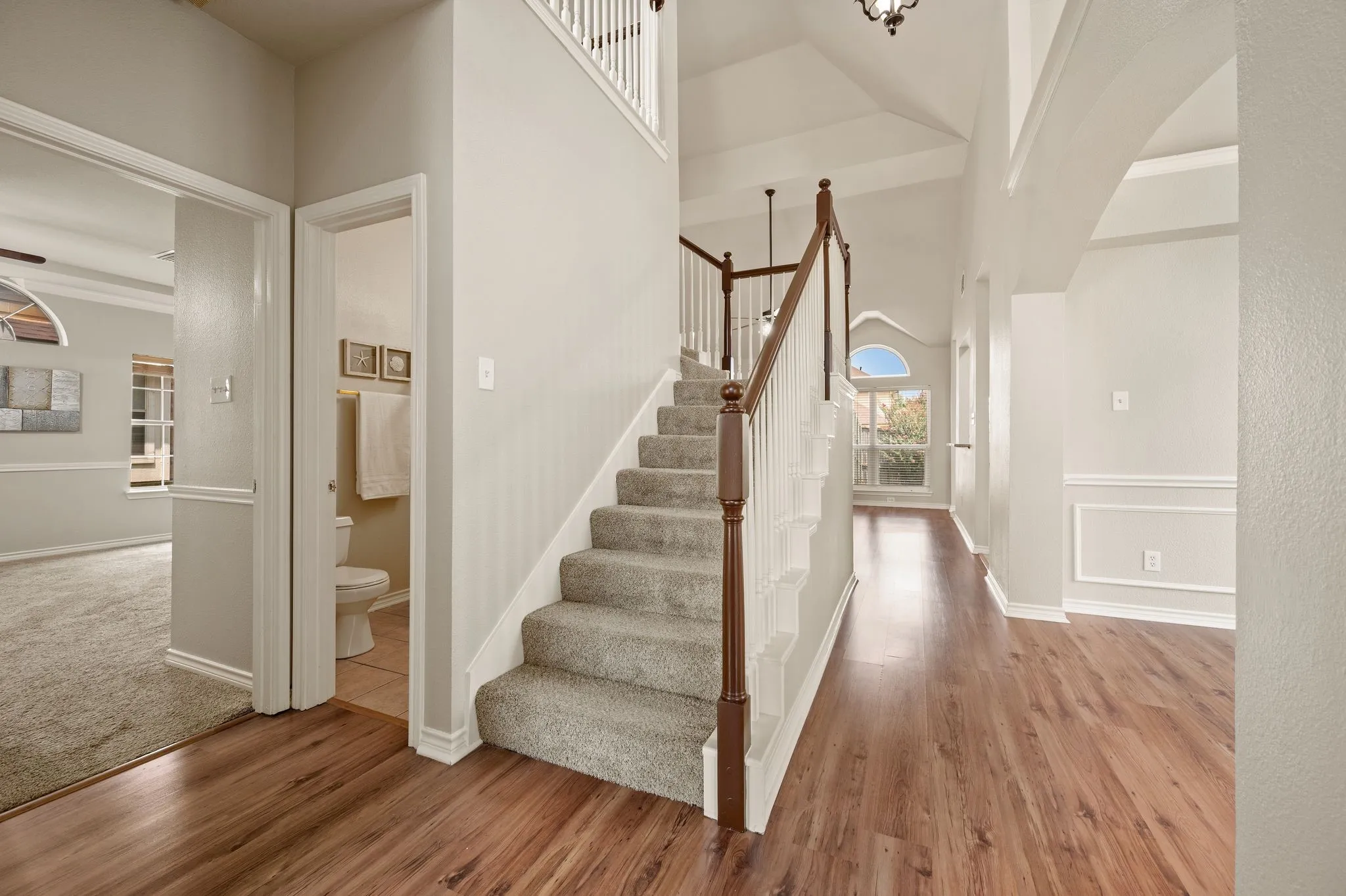 Bright, open foyer showcasing gleaming wood-finish flooring, a elegant staircase with wood railings and metal balusters, soaring ceilings with abundant natural light from upper windows, and crisp white wainscoting and trim throughout