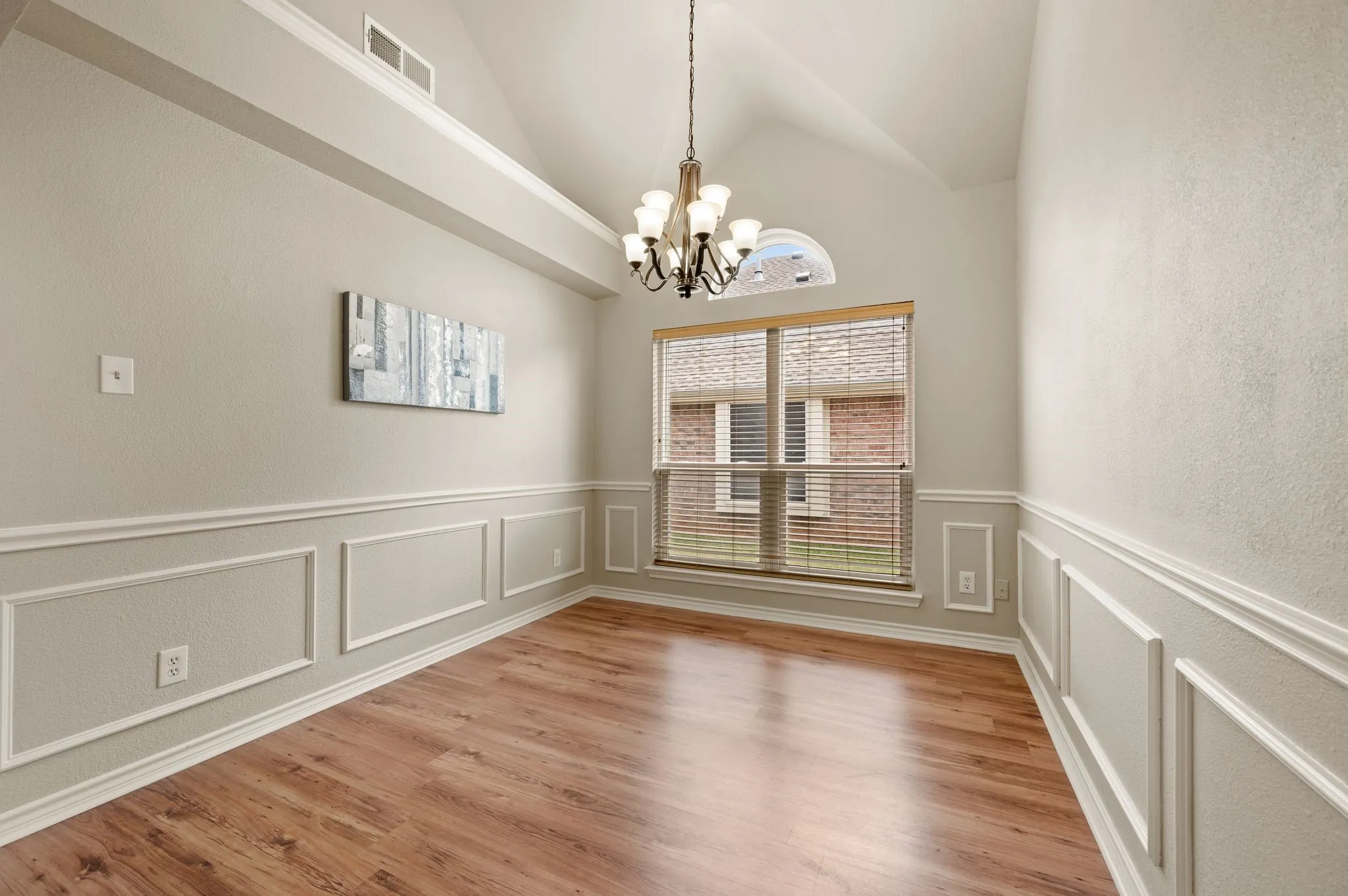 Vaulted ceiling room featuring wood-finish flooring, wainscoting, a traditional chandelier, and a large window with blinds and an arched transom