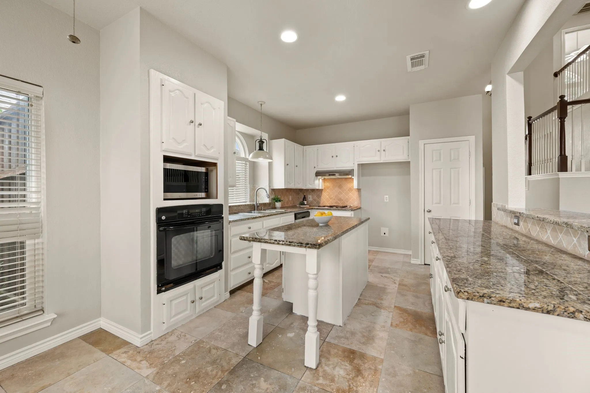 Bright and spacious kitchen featuring white cabinetry, granite countertops, a center island with seating, stainless steel built-in appliances including double wall ovens and microwave, recessed lighting, travertine-finish flooring, and abundant natural light from multiple windows