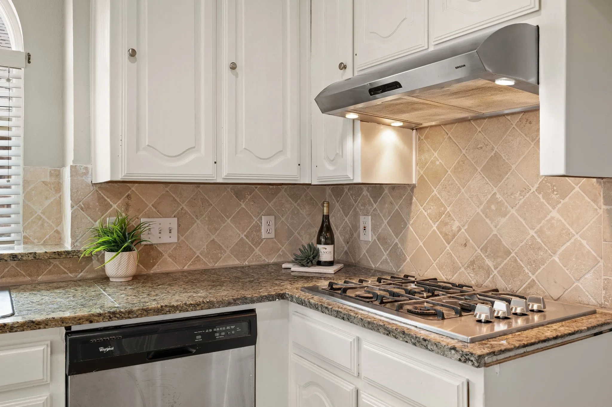 Kitchen featuring white cabinetry, granite countertops, a diamond-patterned tile backsplash, stainless steel gas cooktop, and an undermount range hood with integrated lighting