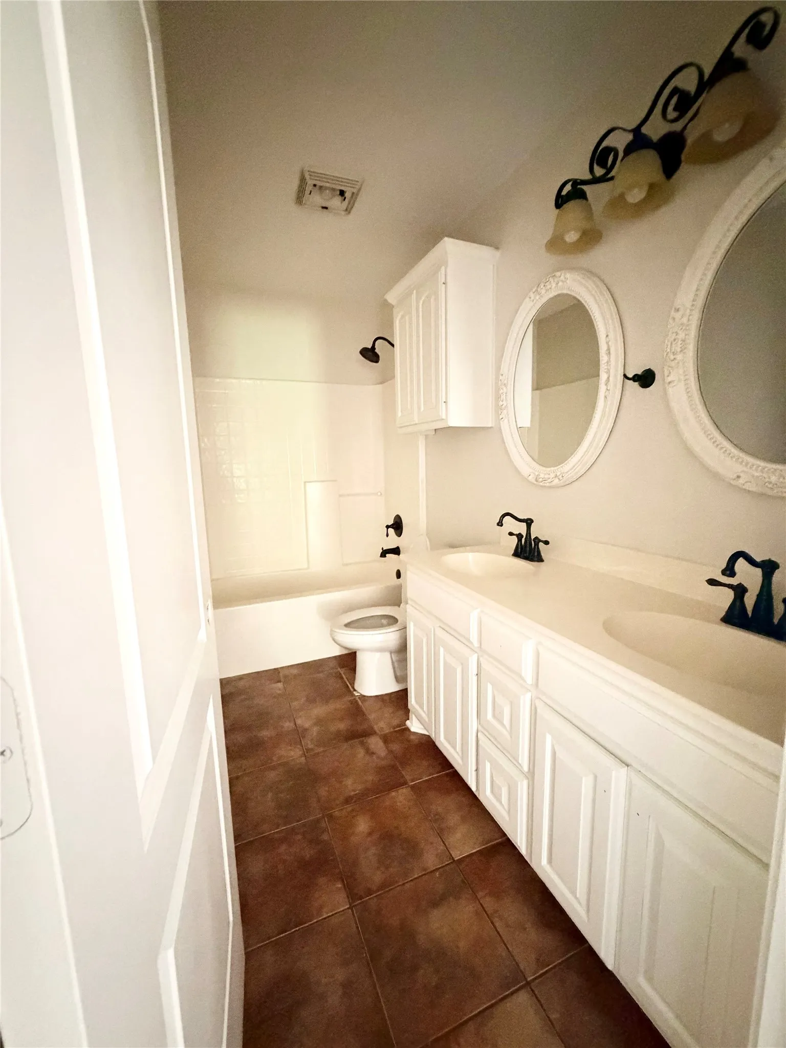 Dual vanity with integrated sinks, white cabinetry, decorative wall sconces, oval mirrors, and brown tile flooring