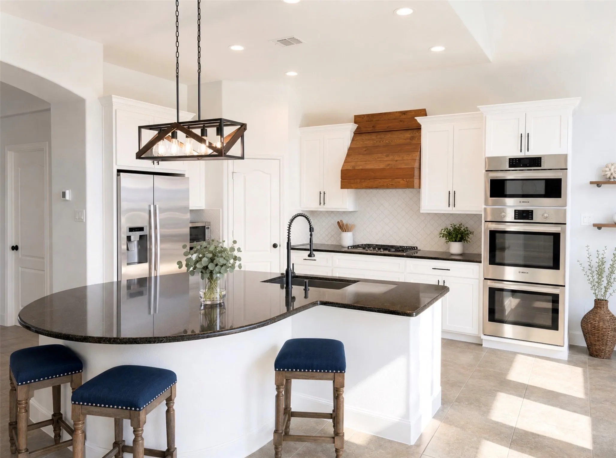 Spacious kitchen featuring an extended island with dark countertop, white cabinetry, integrated stainless steel appliances, a unique wood-finish range hood, and tile flooring