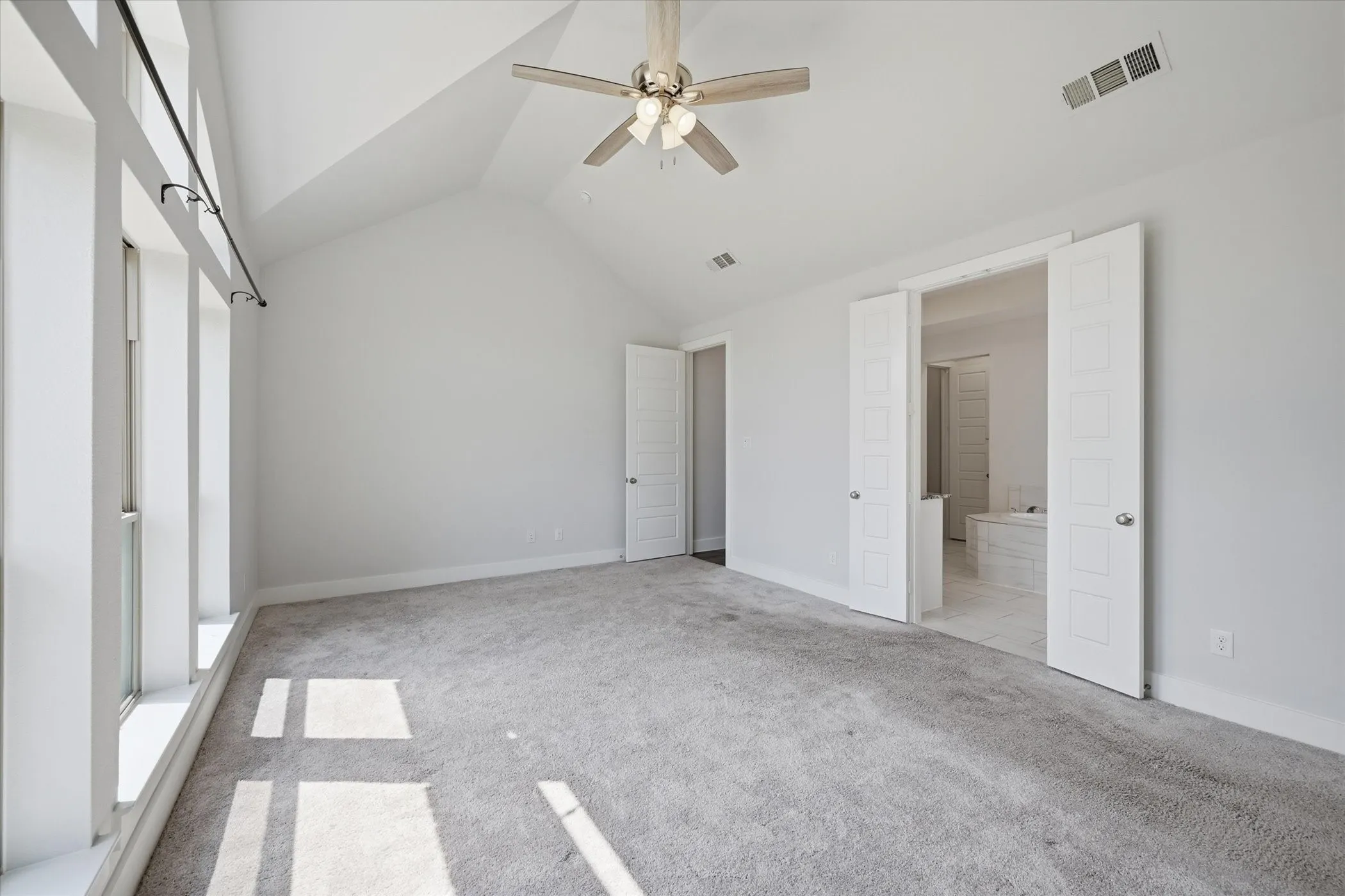 Vaulted ceiling with an attached ceiling fan, large windows with black curtain rods, light grey carpet, and double panel doors leading to a tiled space