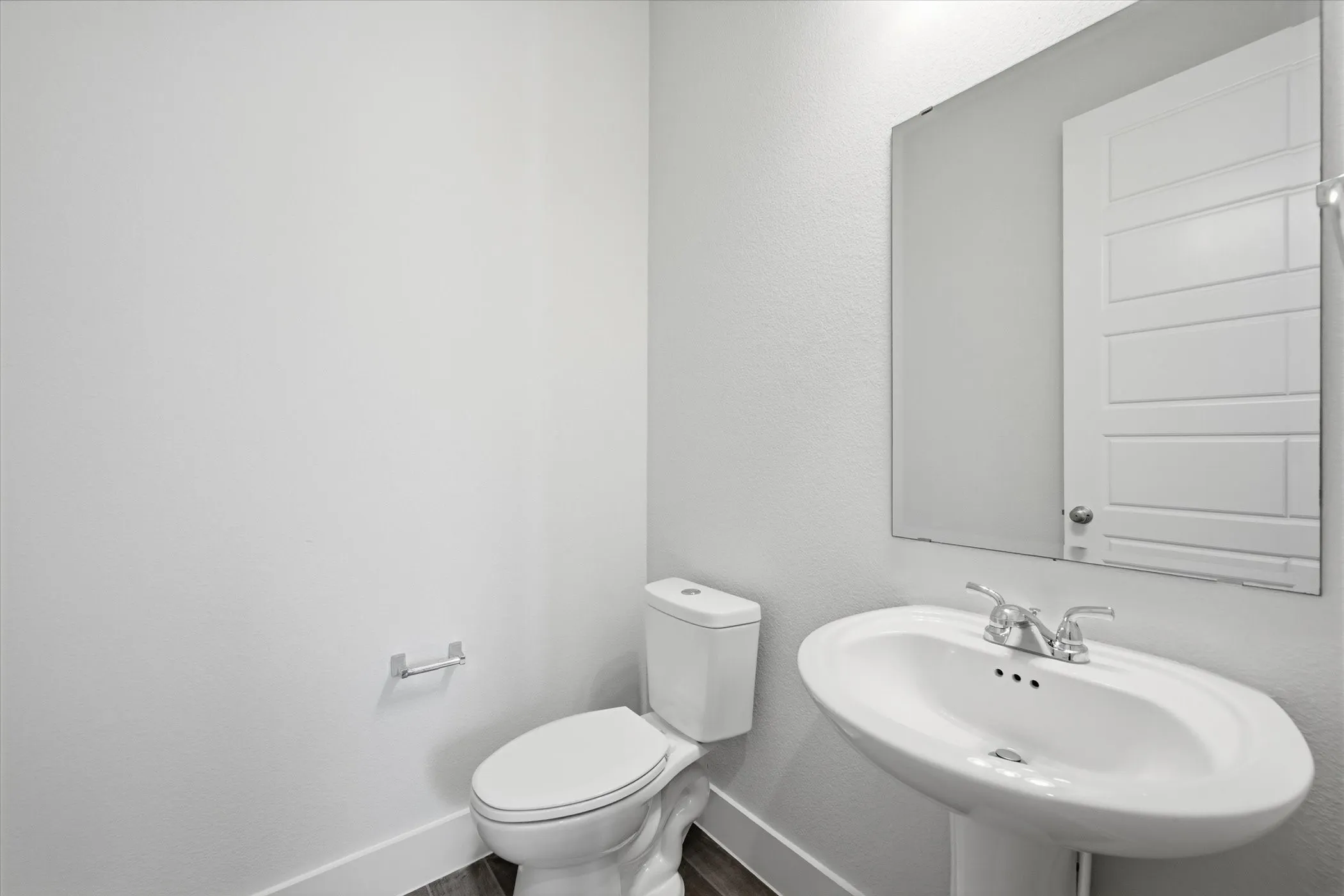 Pedestal sink with chrome fixtures, elongated toilet, wall-mounted mirror, and wood-finish flooring