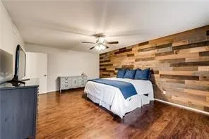 Spacious primary bedroom featuring a striking wood-plank accent wall, hardwood flooring throughout, ceiling fan with integrated lighting, and ample natural light from multiple windows