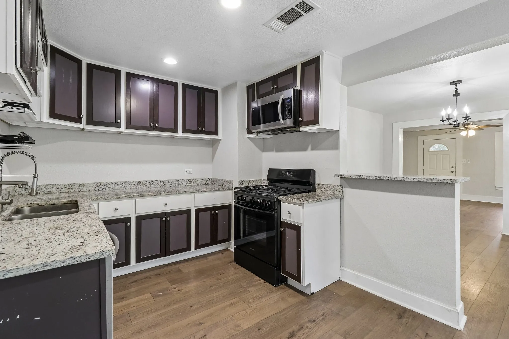 Contemporary kitchen featuring two-tone cabinetry with dark lower units and light upper glass-front doors, granite countertops, stainless steel appliances including a black gas range, recessed ceiling lighting, and wood-finish flooring throughout