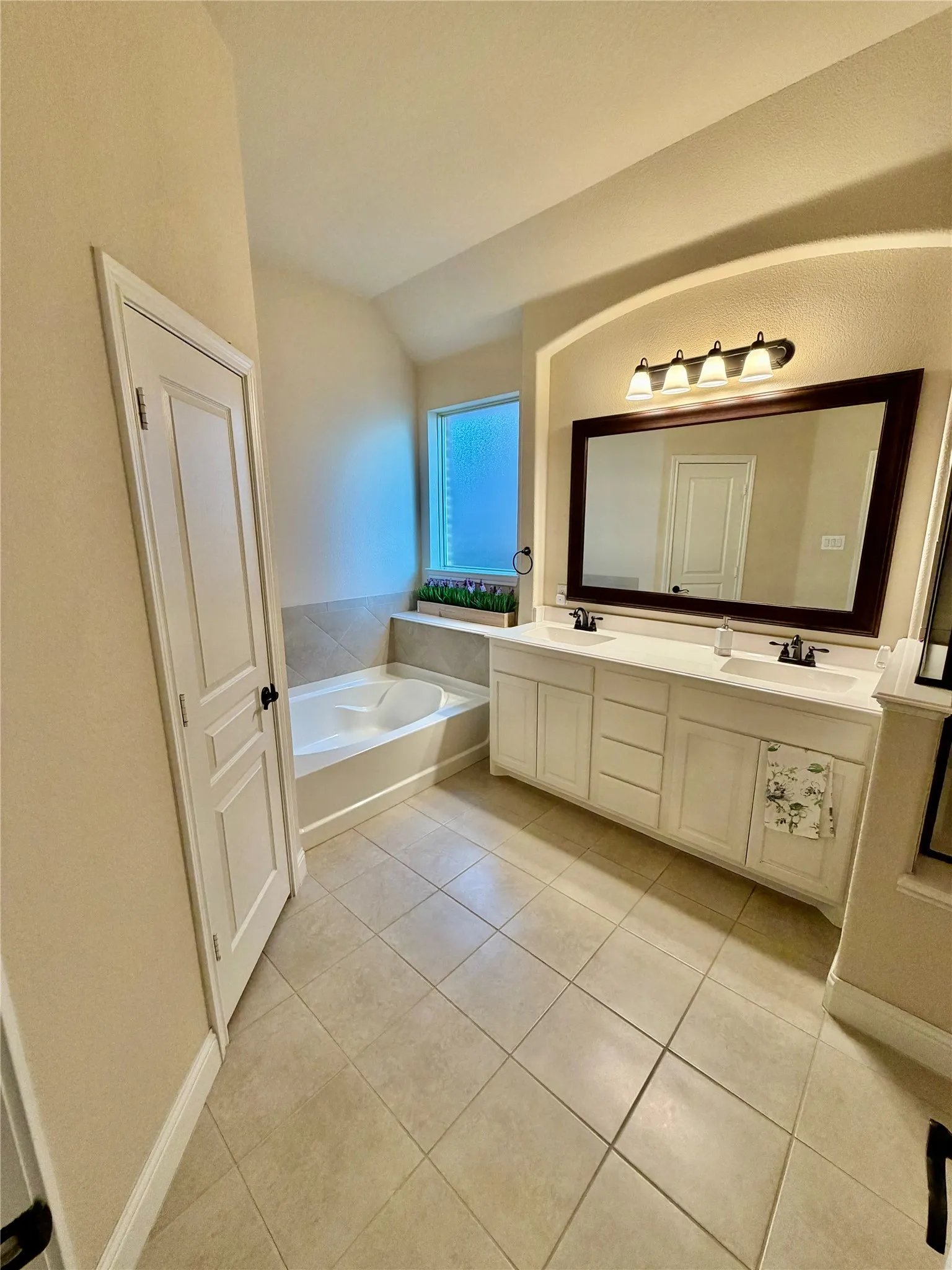 Dual vanity with white cabinetry and integrated sinks, a built-in soaking tub, tile flooring, and a privacy window