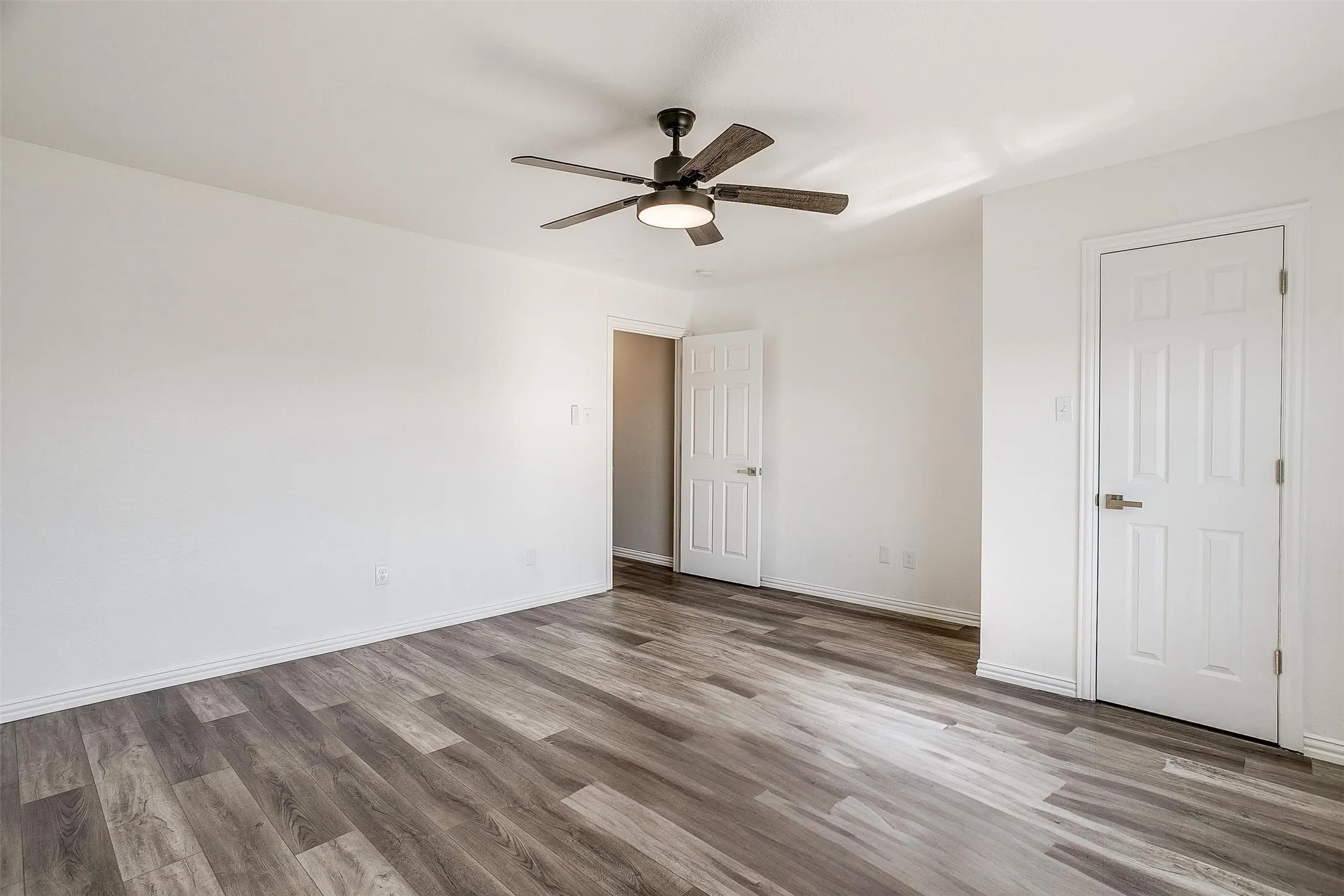 Spacious room featuring wood-finish flooring, white walls, two paneled doors, and a ceiling fan with integrated lighting