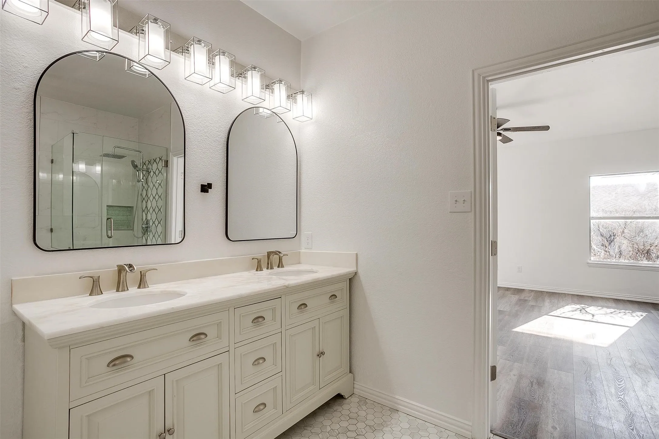 Spa-inspired double vanity bathroom featuring dual arched mirrors with metal frames, linear vanity lighting, cream cabinetry with multiple drawers and hardware accents, and a continuous white countertop