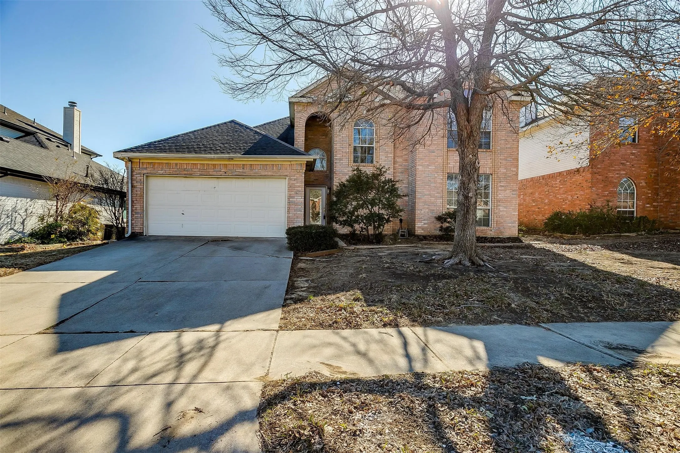 Two-story brick residence featuring a two-car attached garage with white panel doors, circular driveway with asphalt surface, mature shade tree in front landscaping, and multi-pane windows throughout the facade