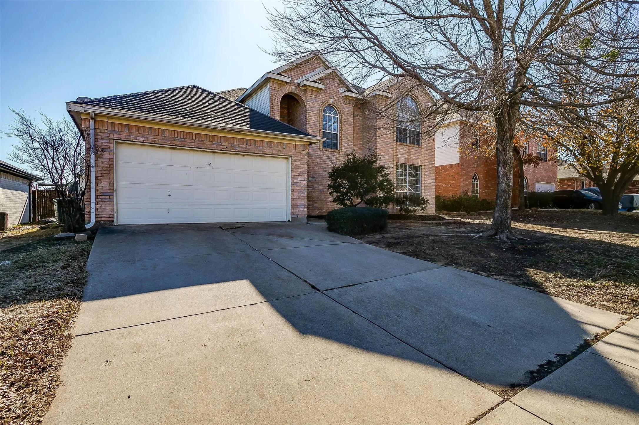 Two-car attached garage with white paneled door, circular driveway with concrete surface, brick exterior with arched architectural details, mature landscaping with established trees, and multi-level roofline with mixed shingle and architectural finishes
