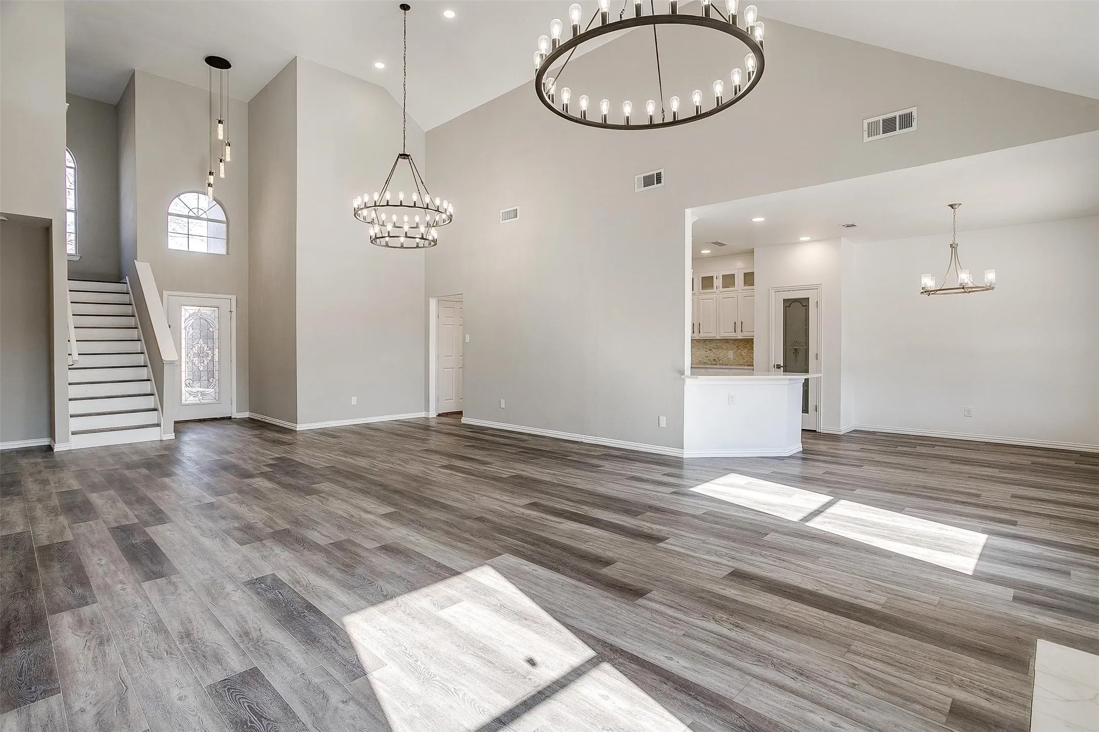 Vaulted ceilings, wood-finish flooring, multiple contemporary chandeliers, and white kitchen cabinetry