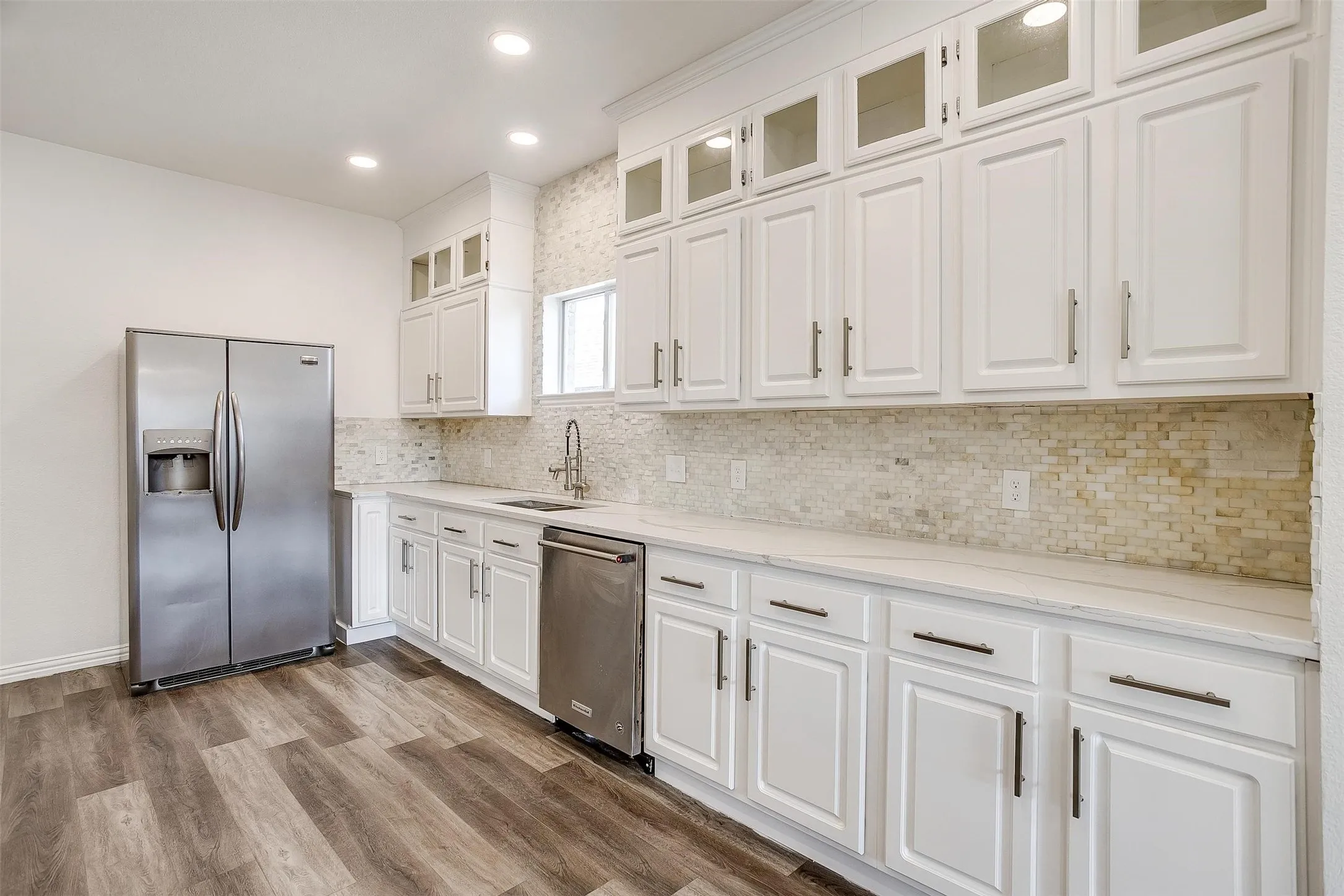 White cabinetry with upper glass-front accents, stainless steel appliances, wood-finish flooring, recessed lighting, and a mosaic tile backsplash